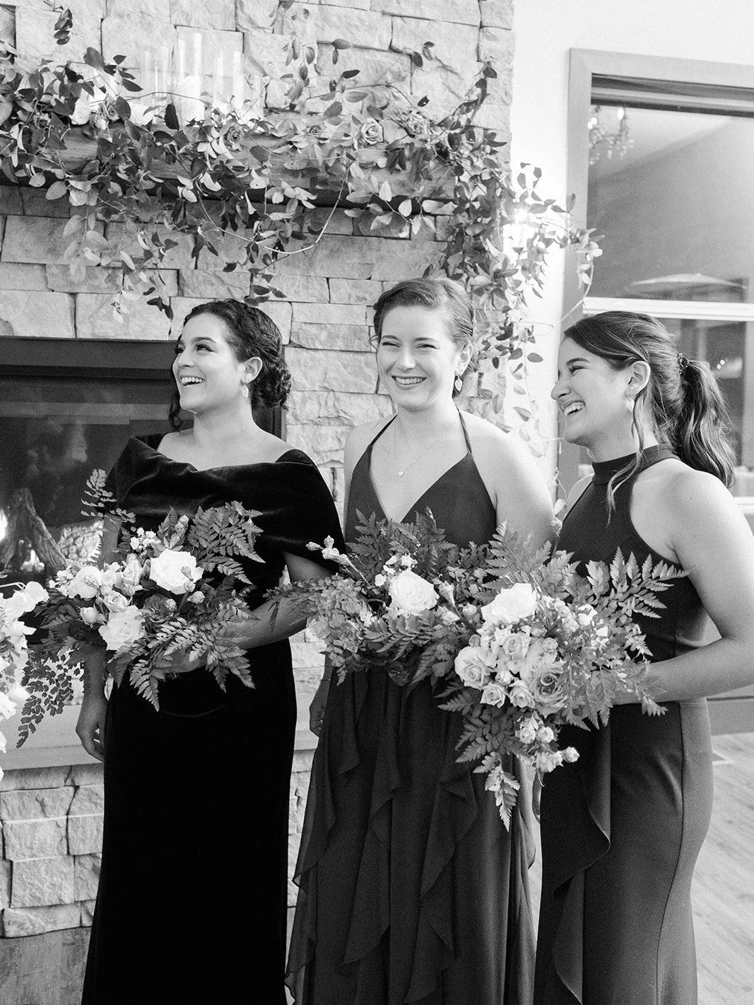 Black-and-white portrait of three attendants holding lush bouquets beside the floral fireplace.