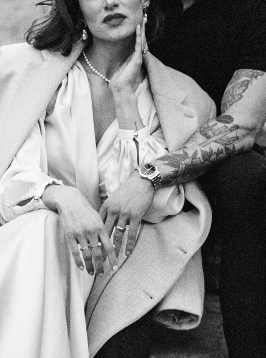 Black and white close-up of a woman in a white dress with pearl jewelry, a tattooed arm wrapped around her.