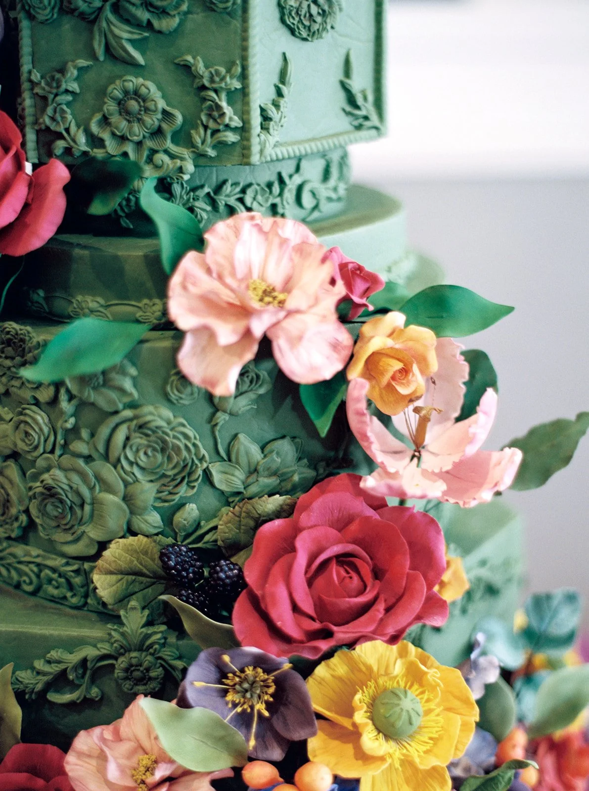 Detailed view of embossed floral patterns on a green tiered cake accented with red and pastel sugar flowers.
