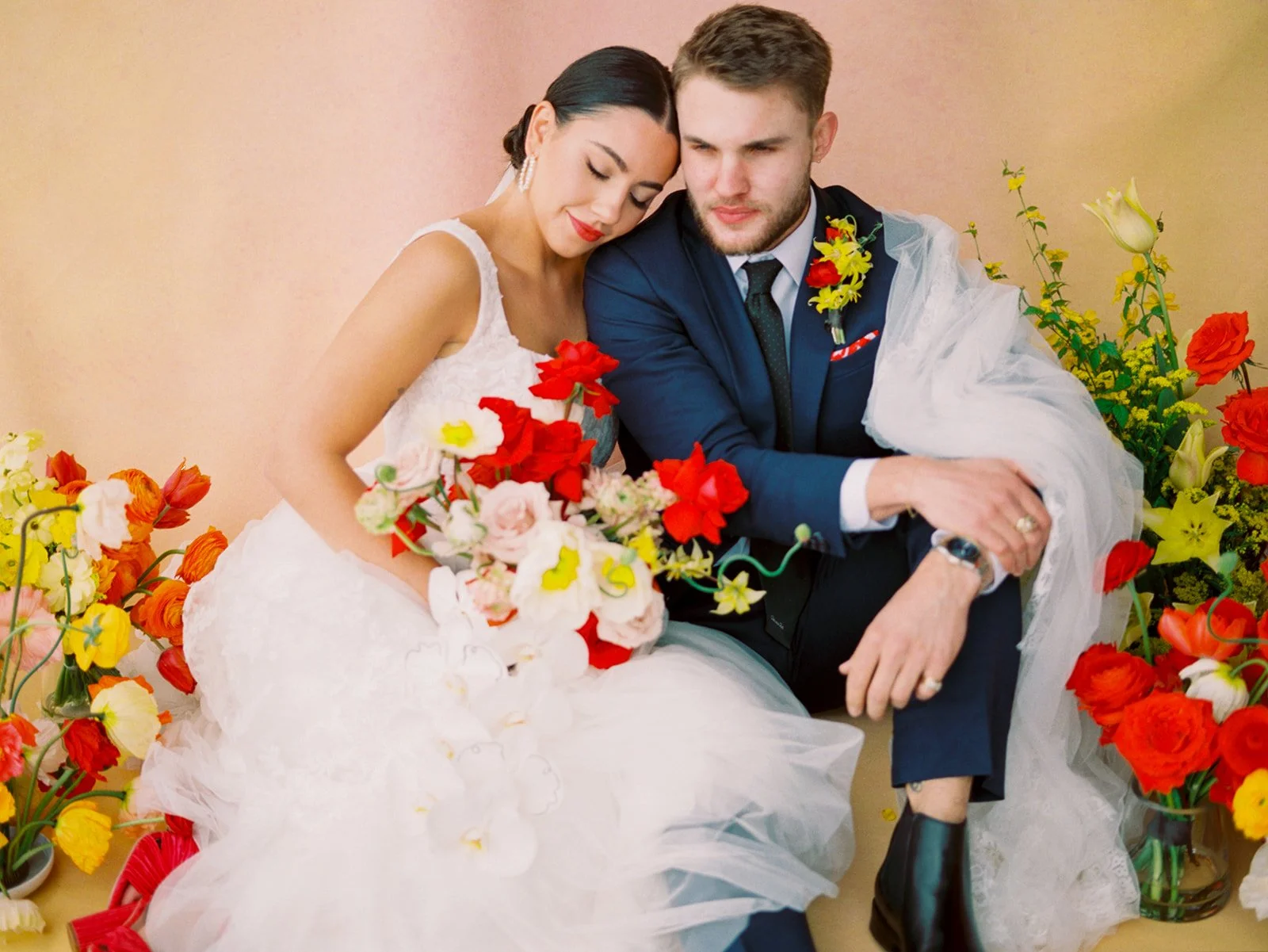 Bride leaning on groom’s shoulder while seated among vibrant floral arrangements