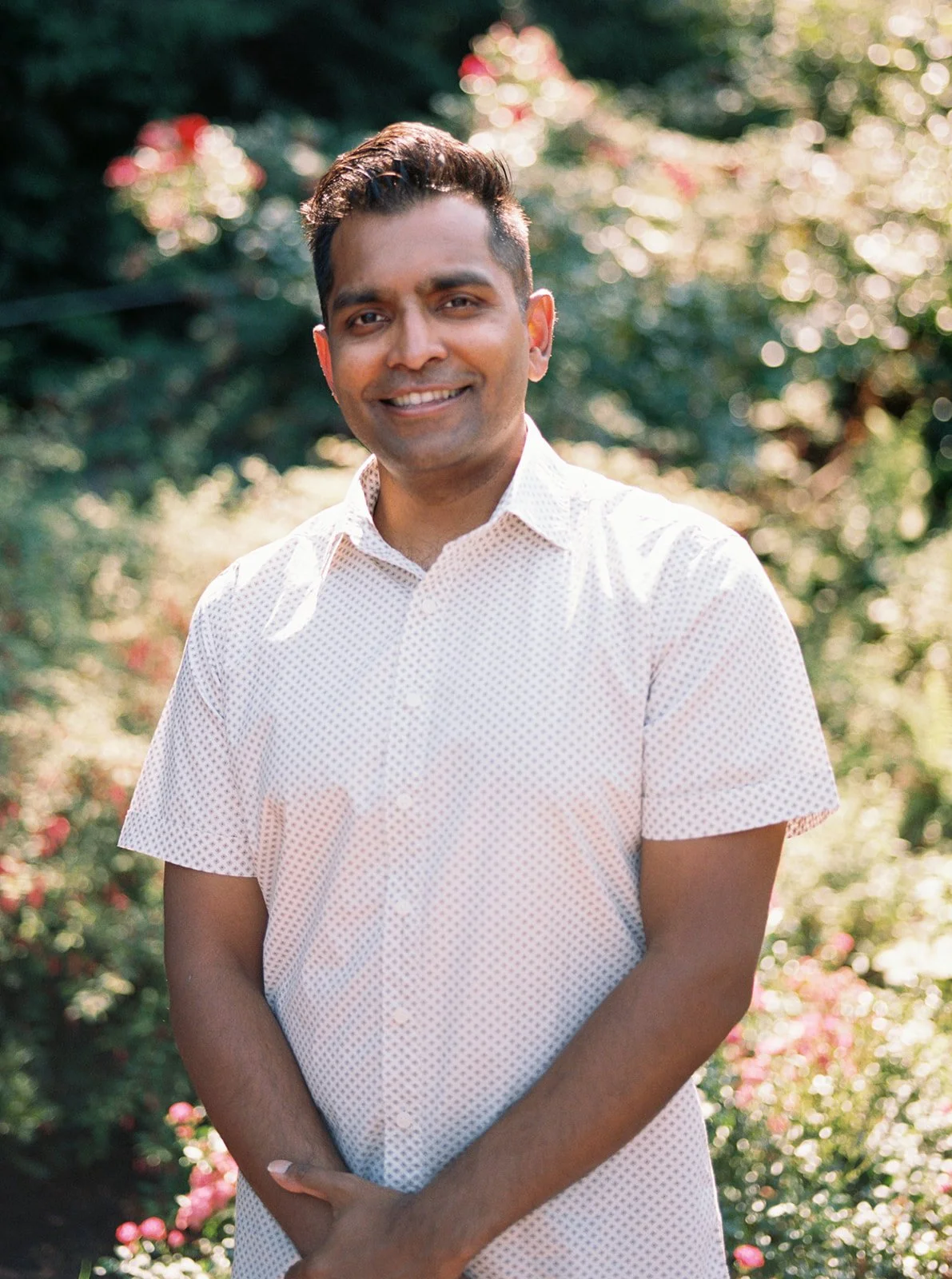 A solo portrait of a man smiling warmly in a white shirt, surrounded by soft bokeh of pink roses.