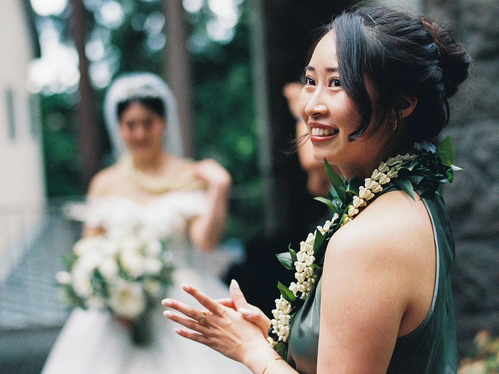 Close-up of a bridesmaid in green wearing a white floral lei laughing with hands clasped, the bride in her veil softly blurred in the background