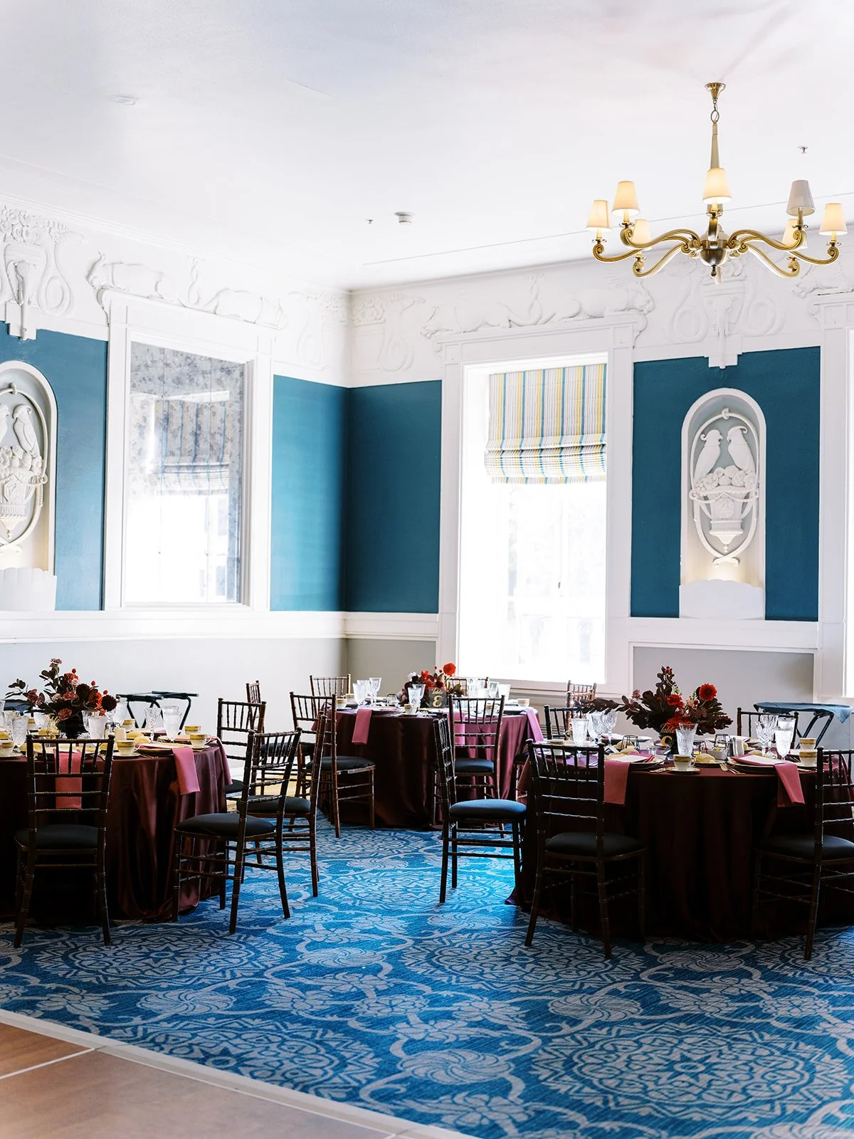 Bright reception room with patterned blue carpet, chandeliers, and tables styled with burgundy linens and florals.