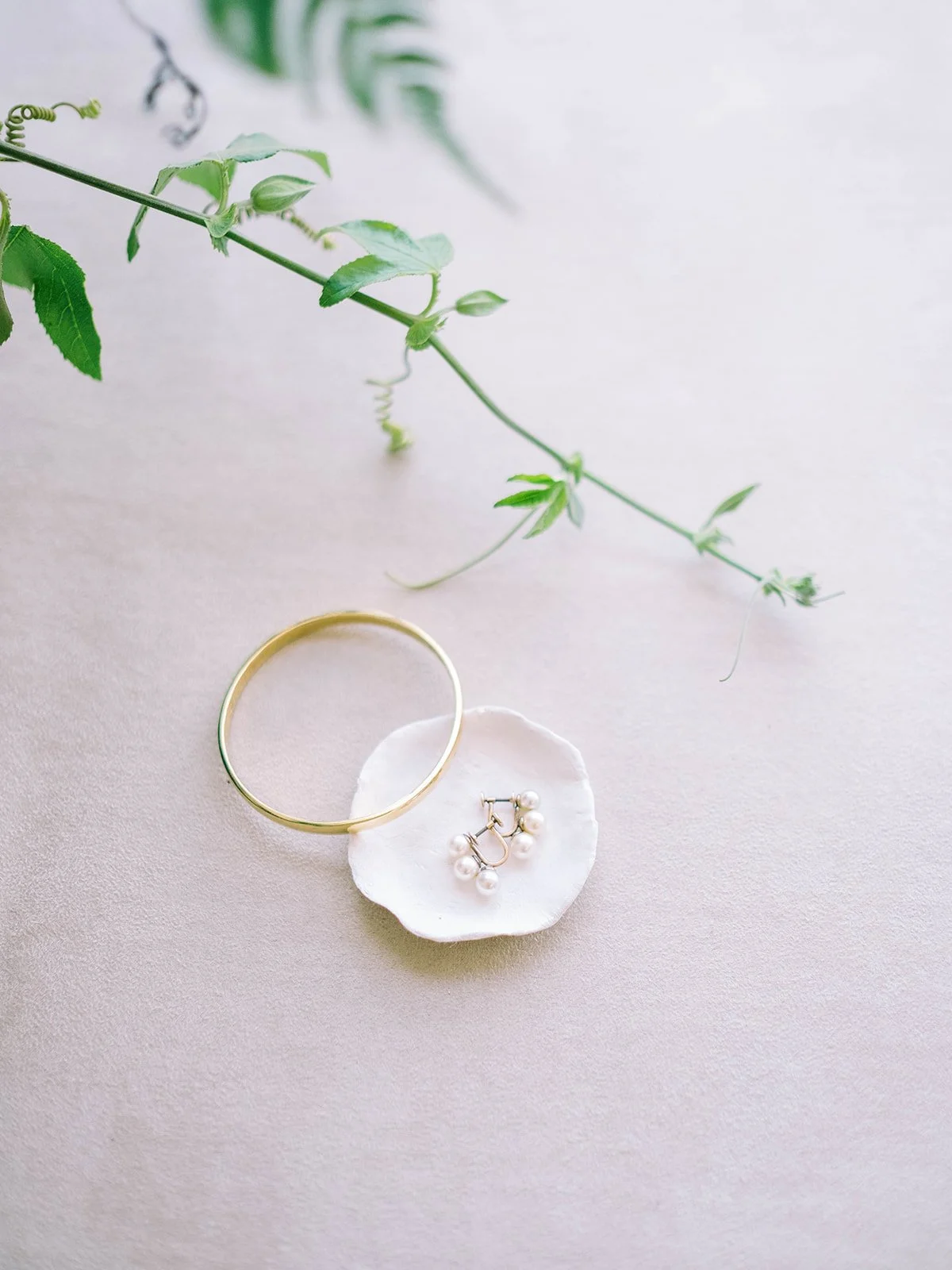 Gold bracelet and pearl earrings arranged with a trailing green vine on a pale surface.
