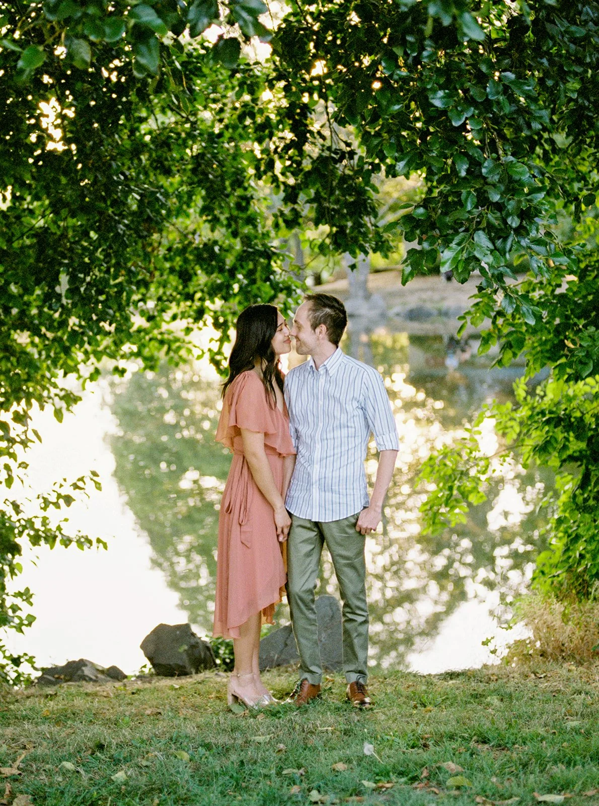 Couple standing under trees by the lake, soft light filtering through leaves in Oregon