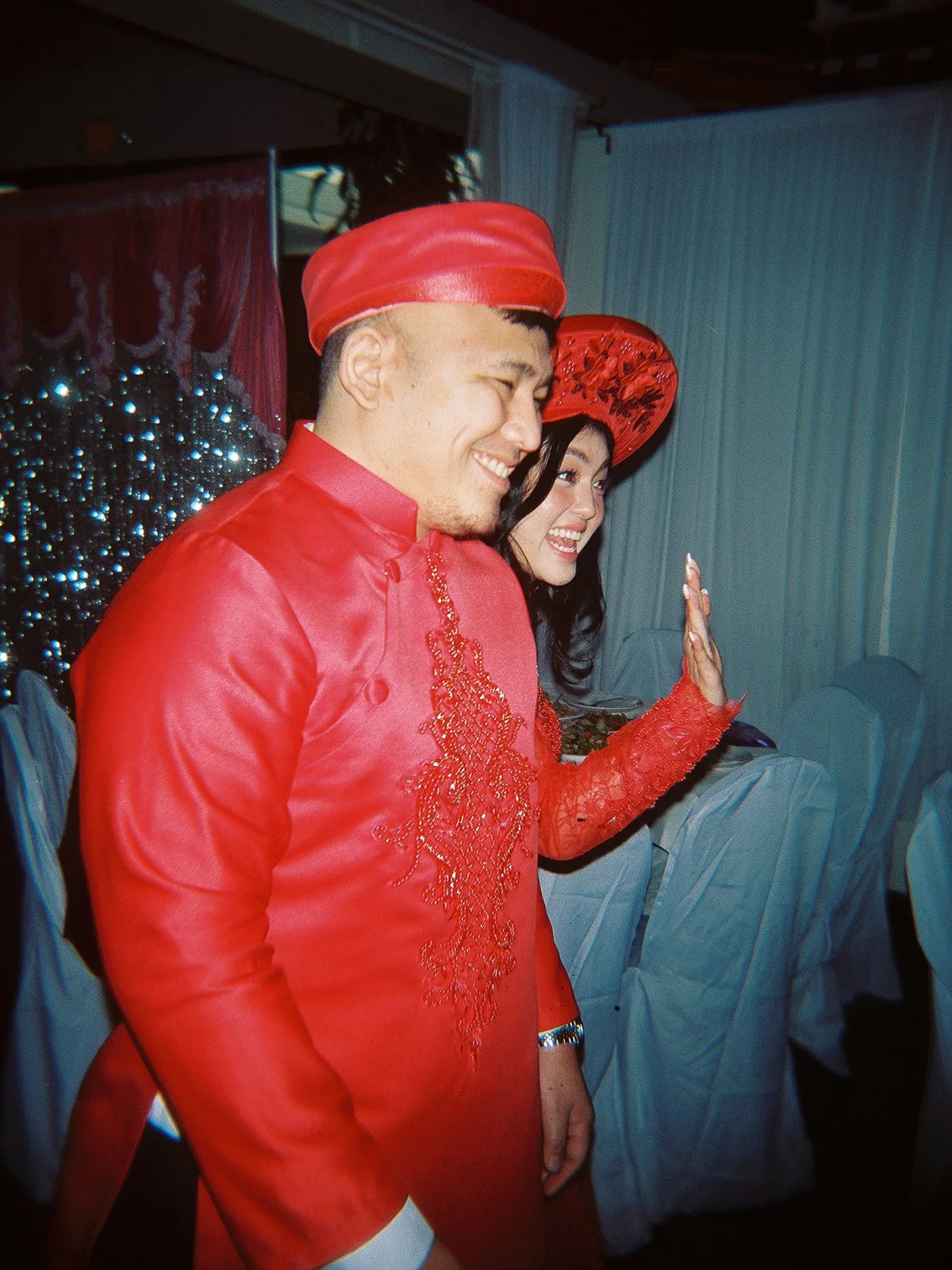 Bride and groom laughing together at the reception in red embroidered Vietnamese áo dài, she wearing a red nón lá, a sequined backdrop behind them