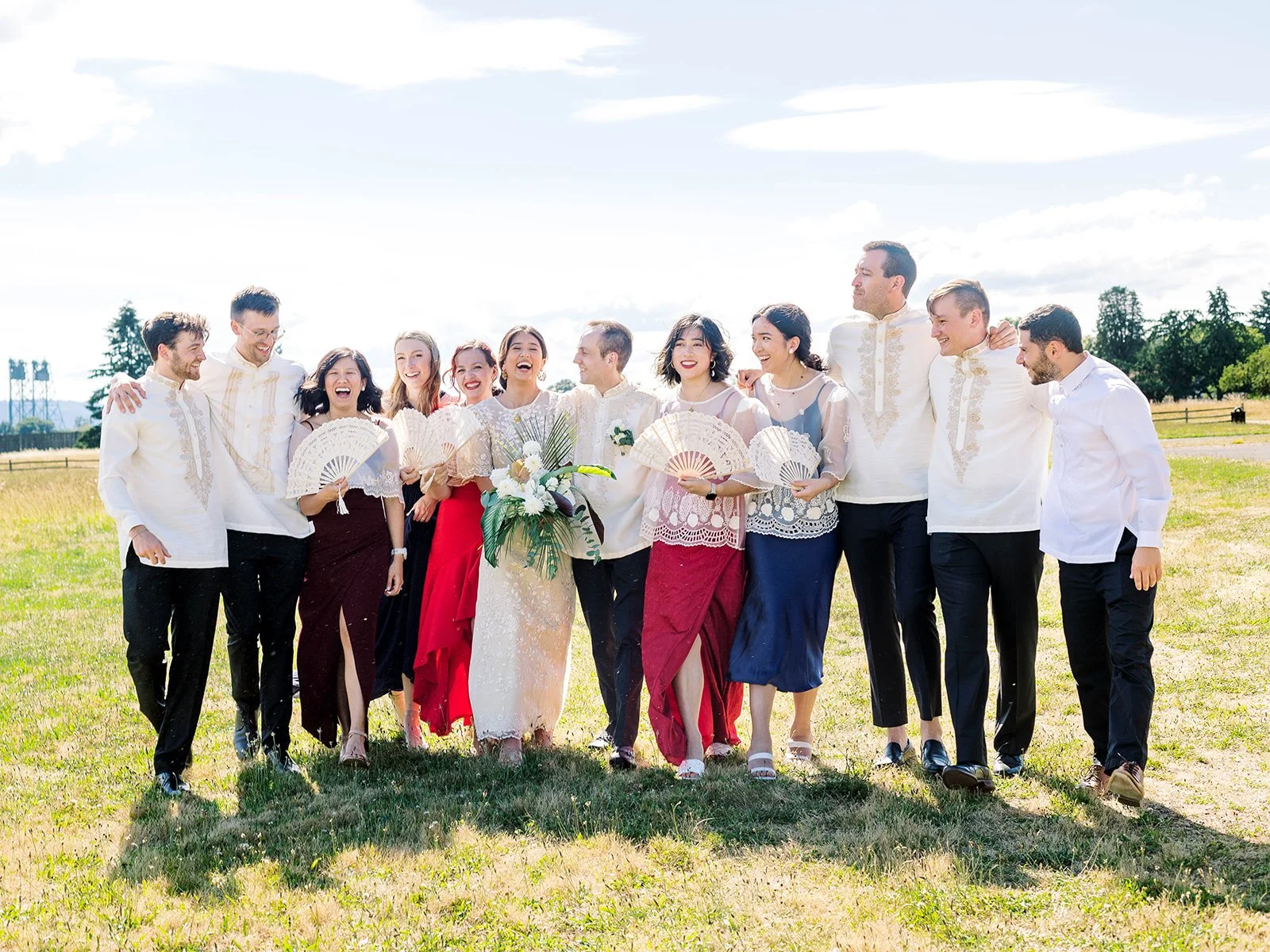 Full wedding party laughing and celebrating together in a sunny grass field, wearing Filipino traditional attire and holding lace hand fans
