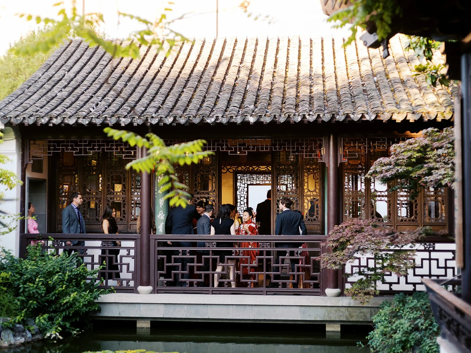 Wedding guests mingle on the covered pavilion terrace, viewed from across the garden through ornate dark wood lattice screens beneath a grey tiled roof.