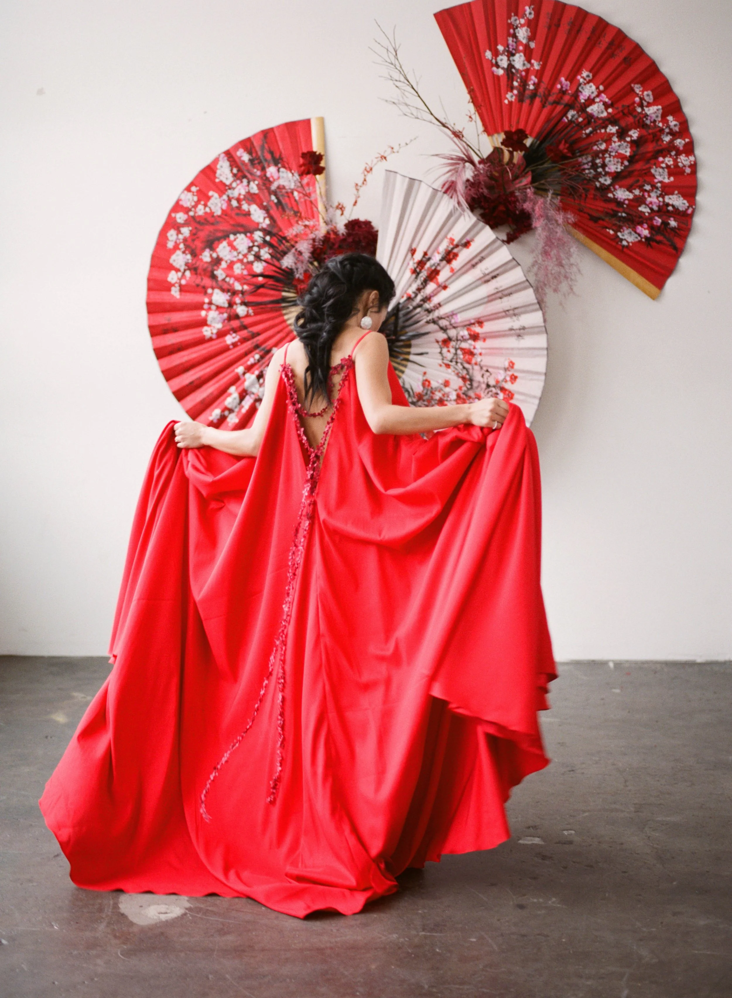 Back view of a flowing red gown with floral detailing framed by oversized Chinese fans.
