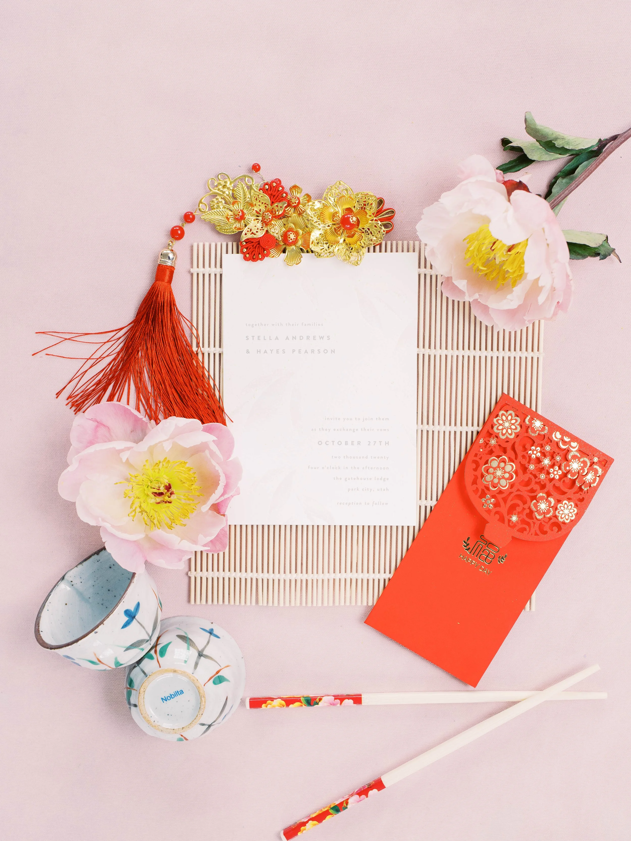 Wedding invitation styled with red envelopes, a gold hairpiece, teacups, chopsticks and pink flowers.