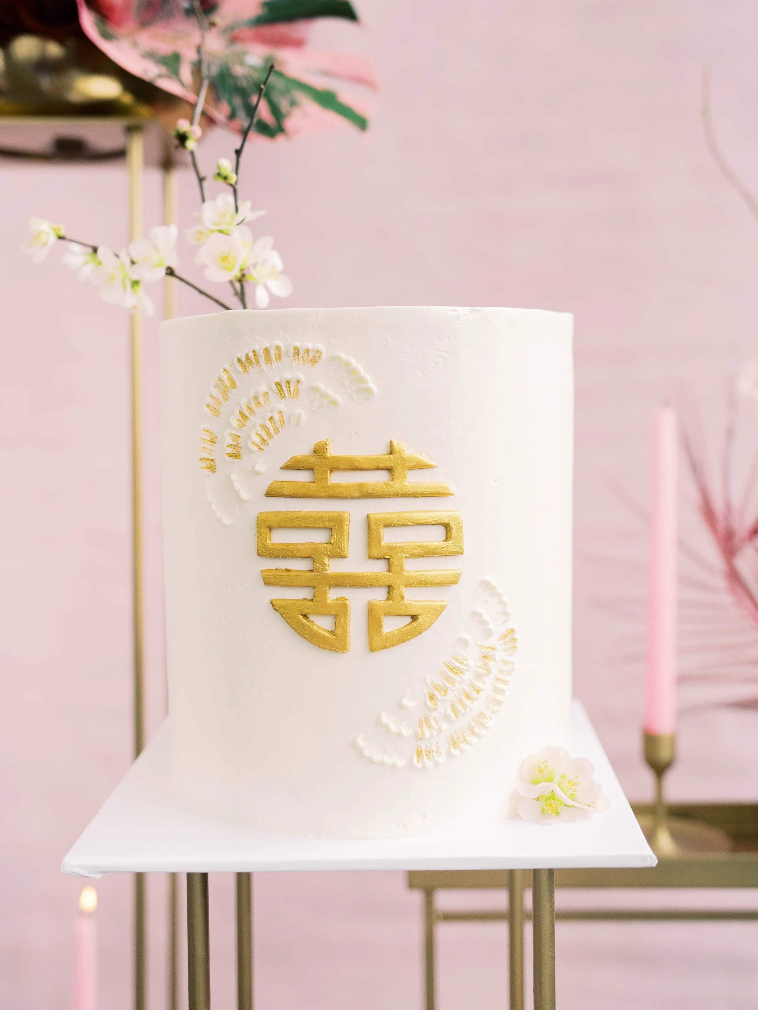 White wedding cake decorated with a gold double happiness symbol against a soft pink backdrop.
