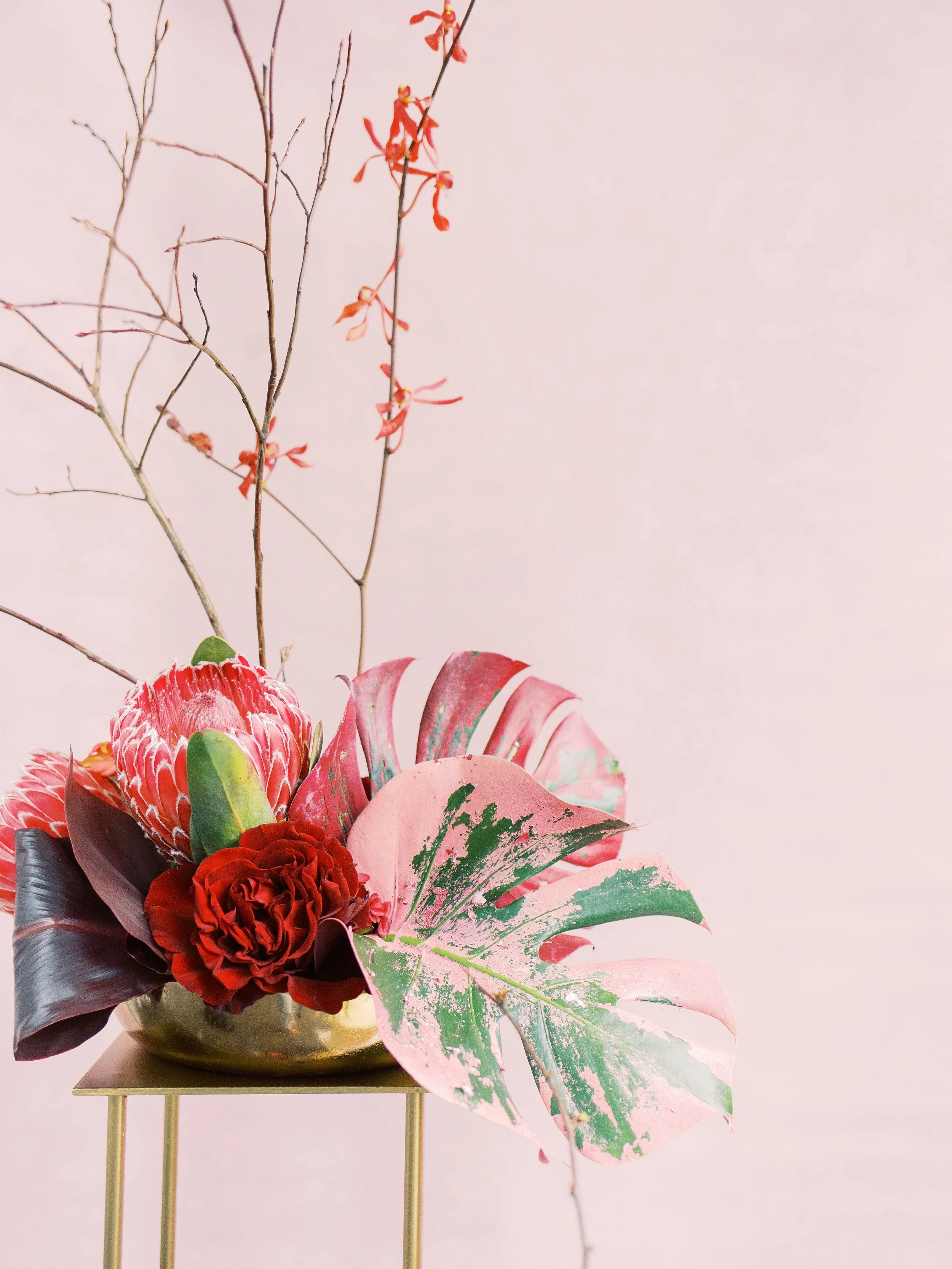 Sculptural red and pink floral arrangement with painted monstera leaves against a blush backdrop.
