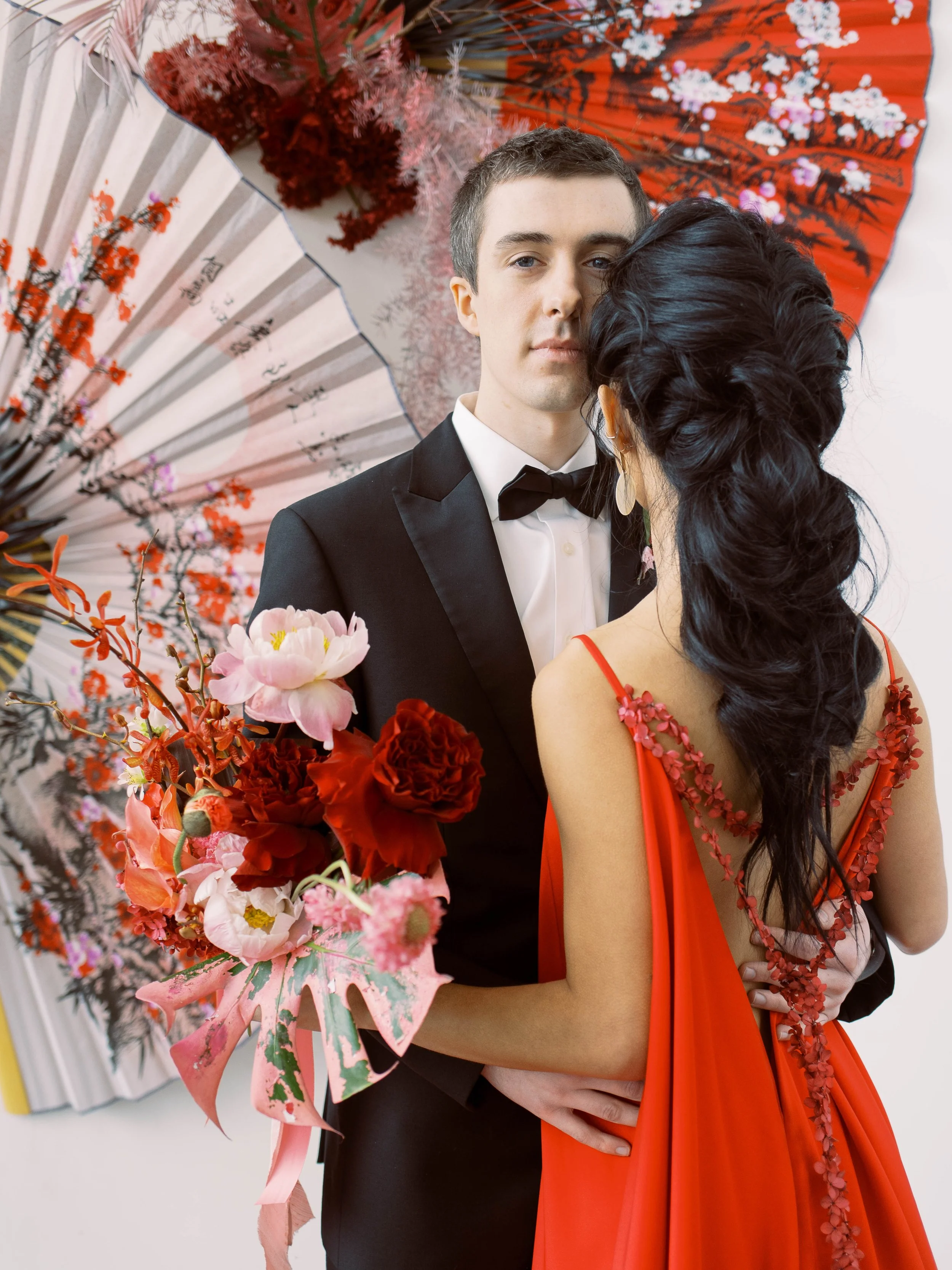 Couple poses closely in front of red and white fans, with a vibrant floral bouquet in the foreground.