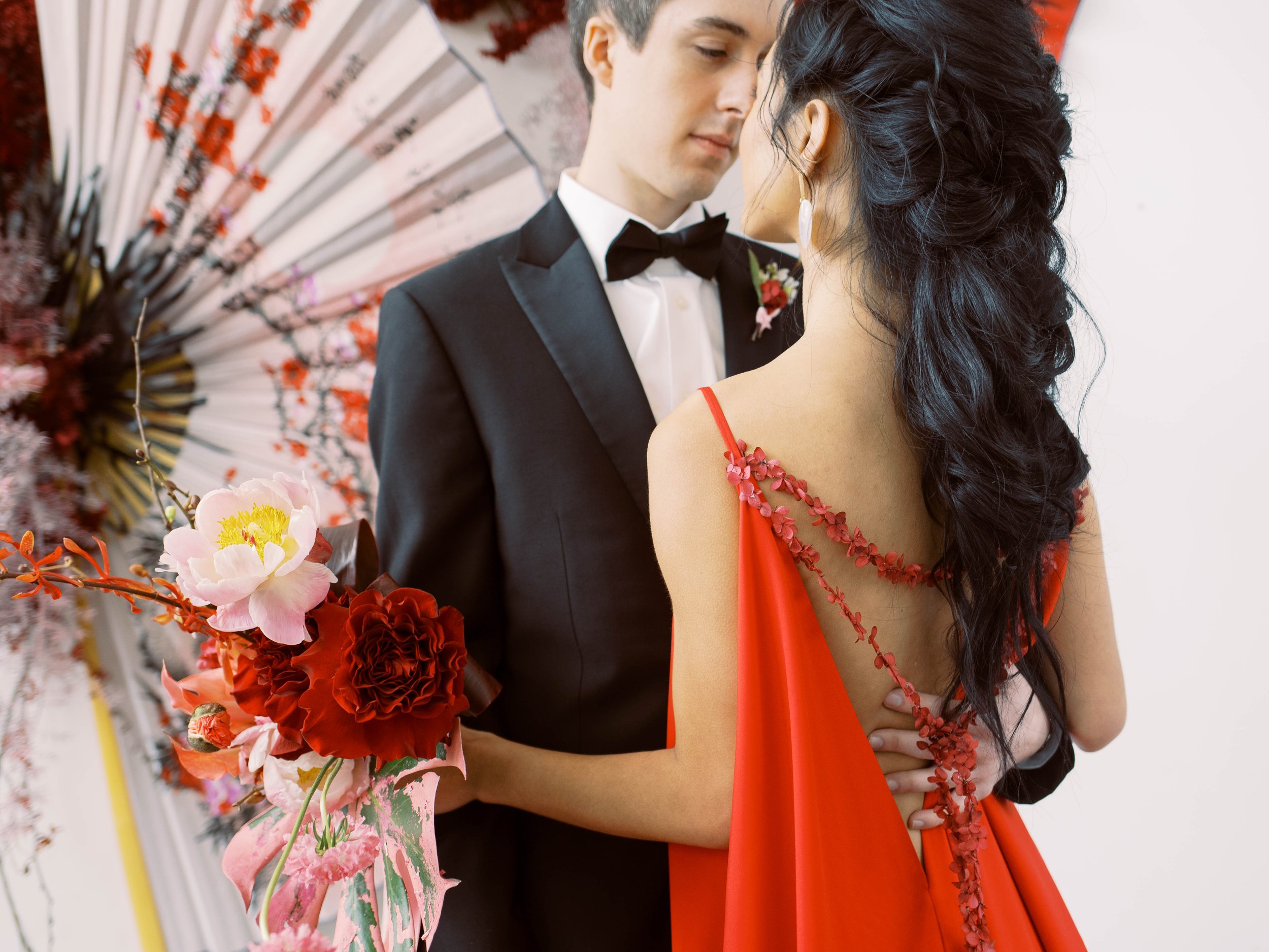 Couple stands close together, highlighting the floral back detail of the woman’s flowing red gown.