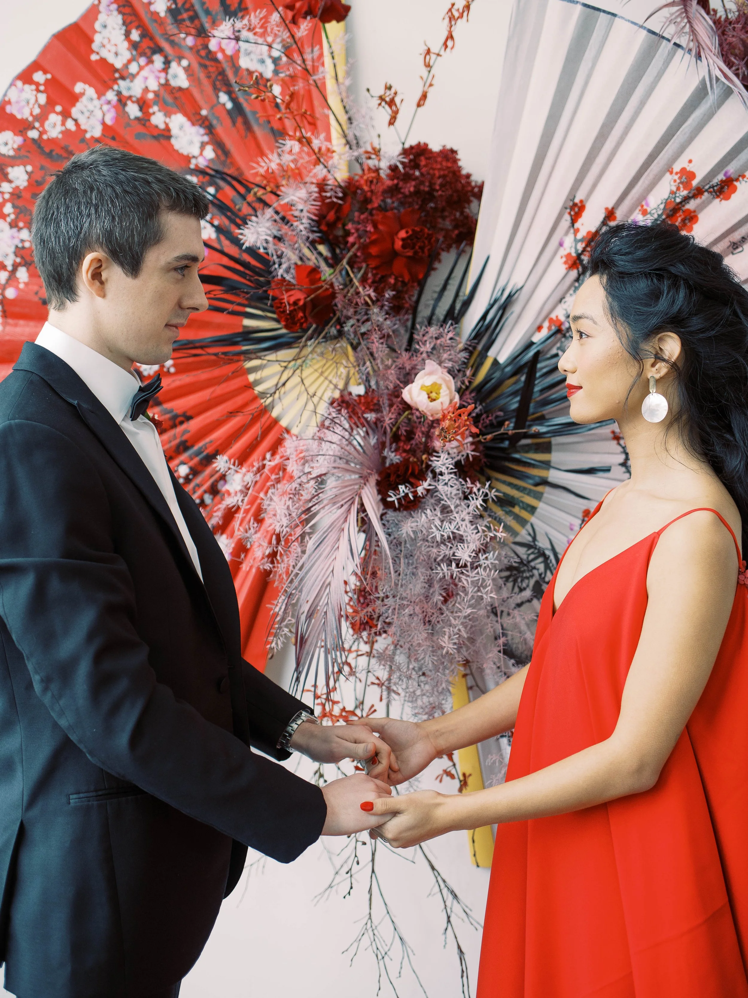 Couple holds hands in front of a floral installation of oversized red and white Chinese fans.