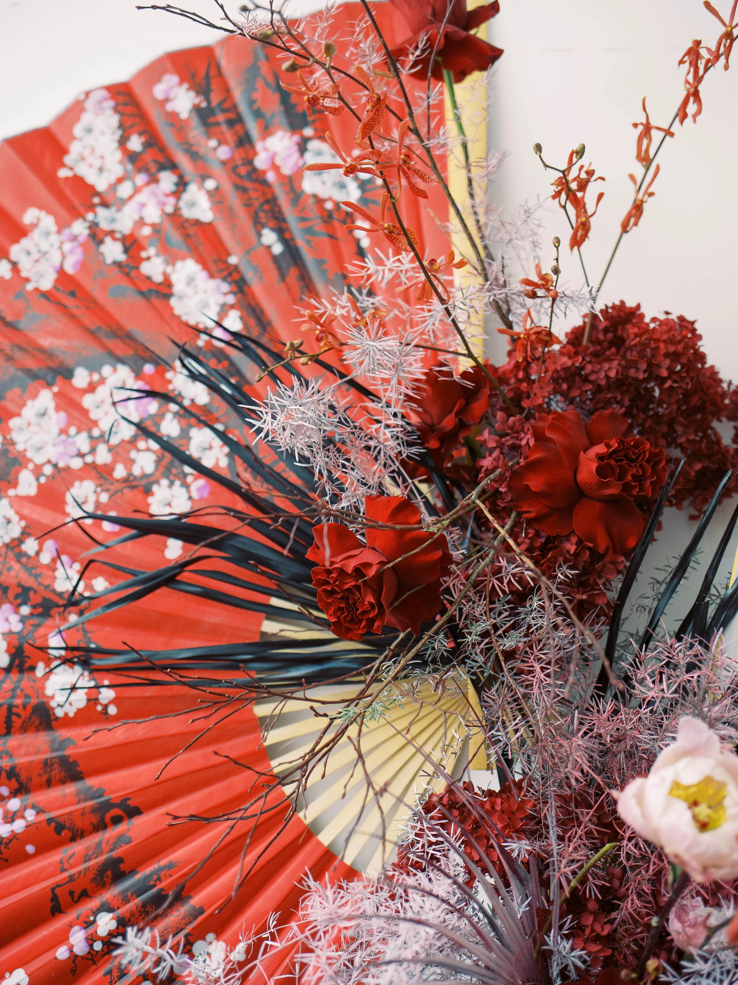 Red roses, pink foliage and delicate branches arranged across a painted Chinese fan.