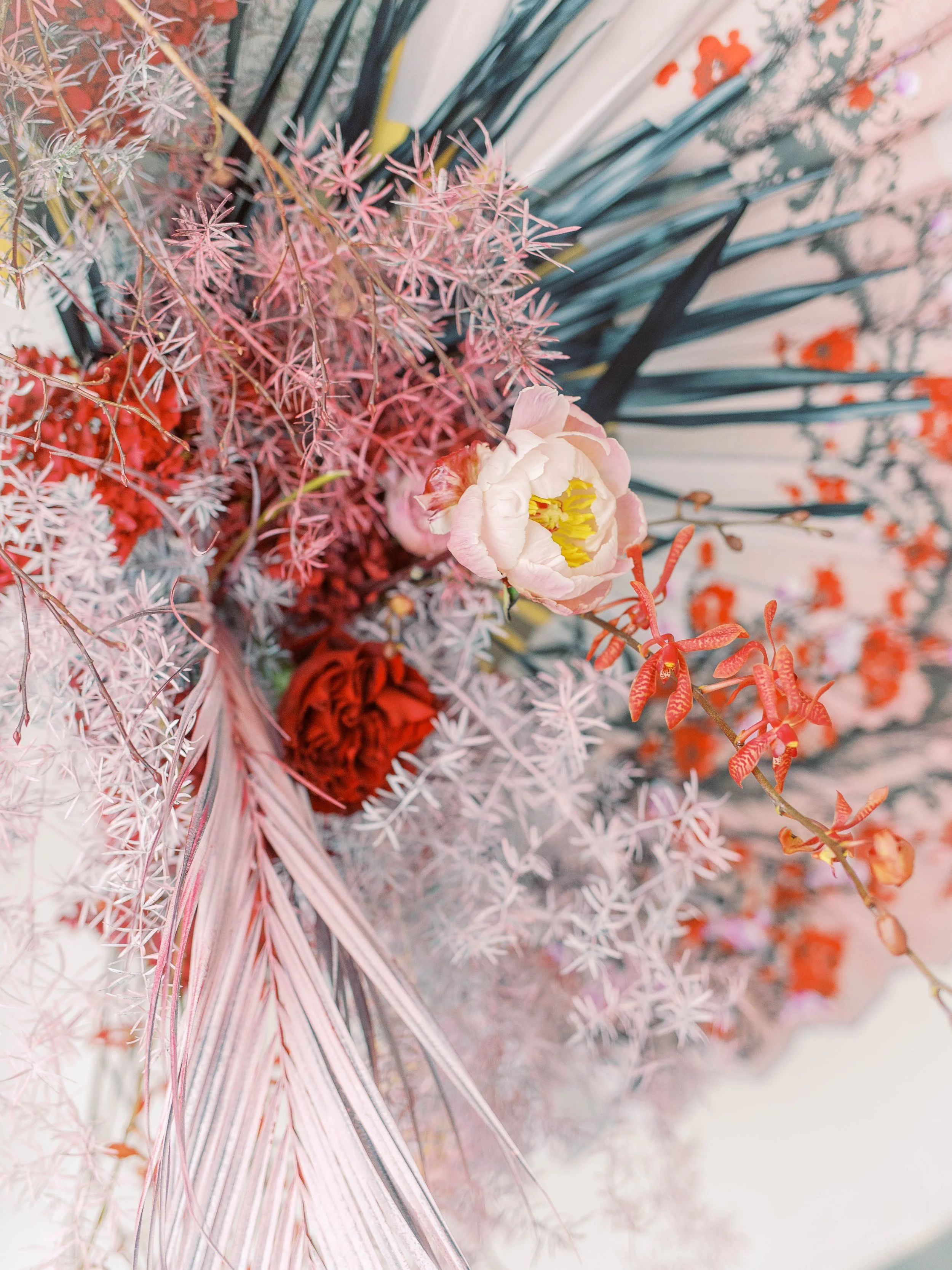 Pale pink peony surrounded by red flowers and airy foliage against a painted fan.