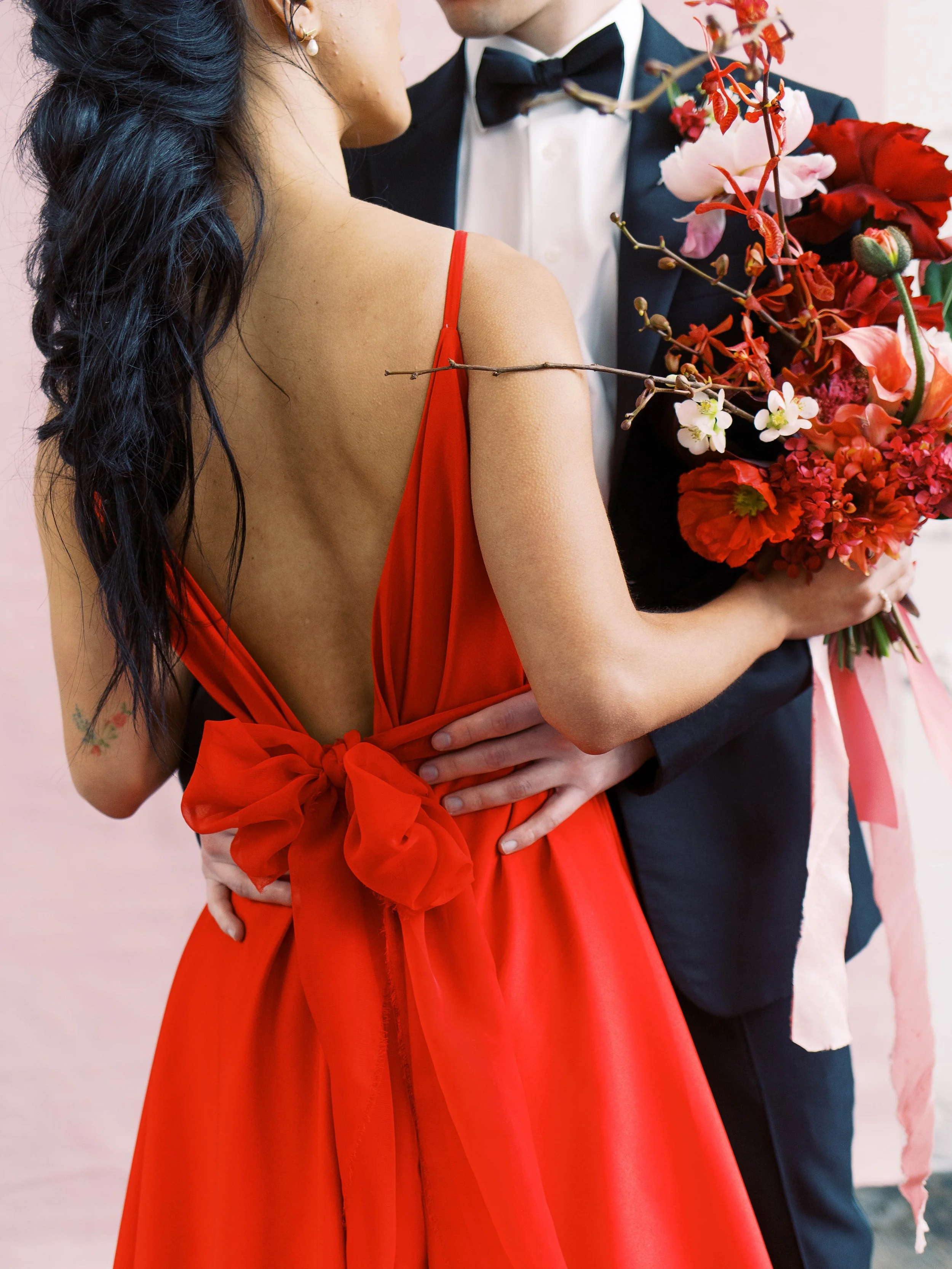 Red gown tied with a large bow as the couple poses together holding a colorful bouquet