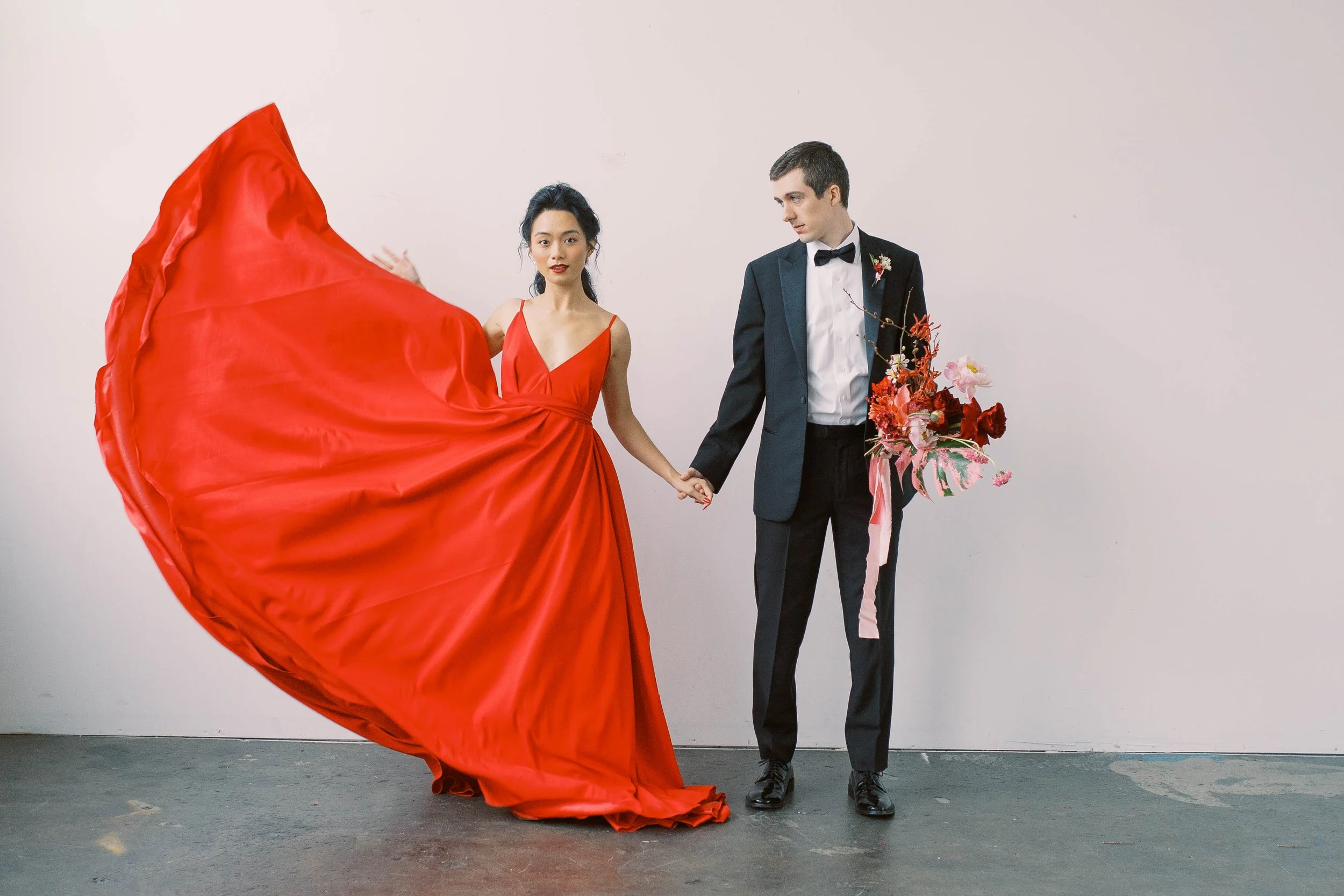 Couple holds hands as the woman’s dramatic red gown billows outward against a pale backdrop.