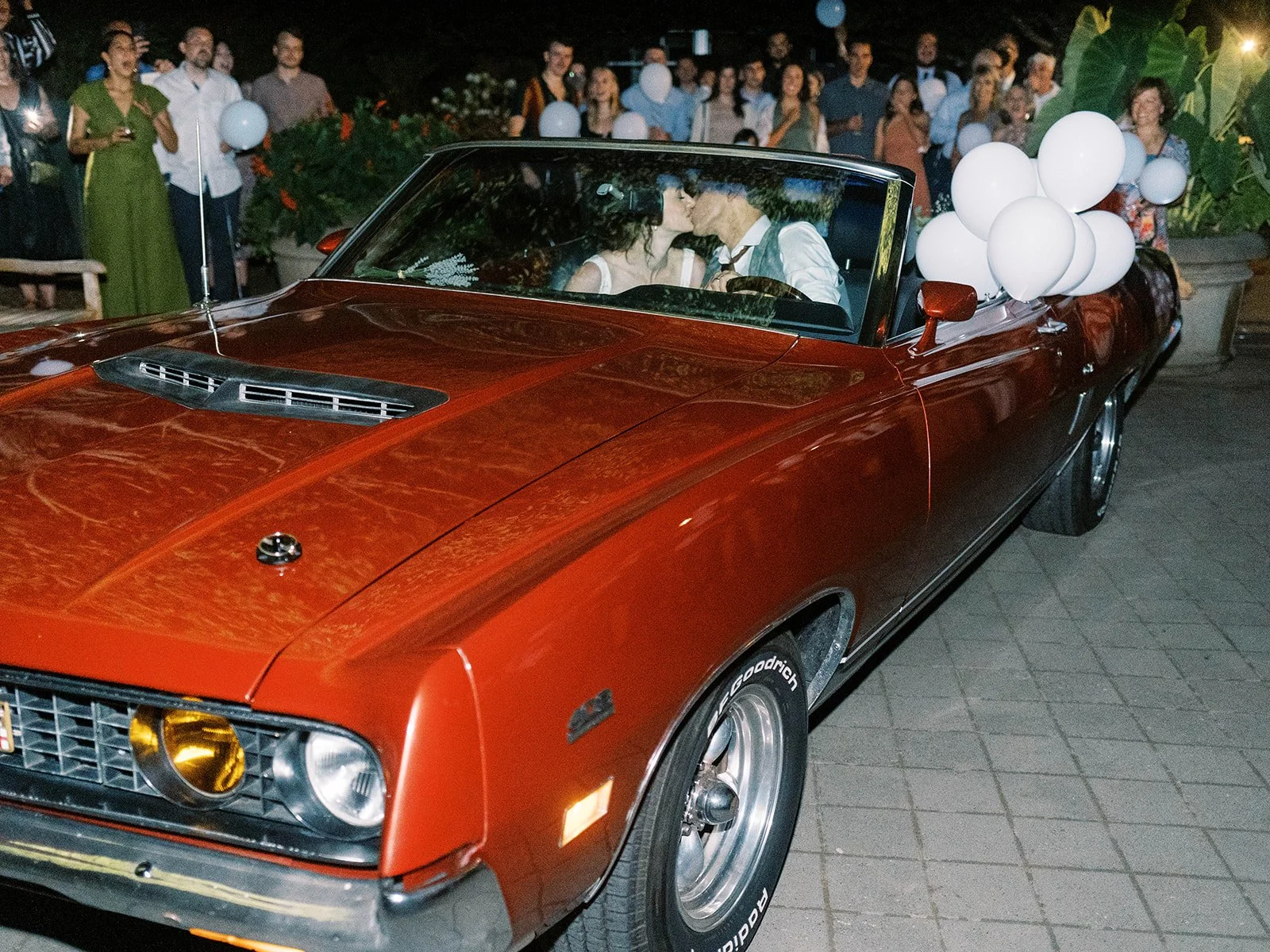 Bride and groom kissing inside a red vintage convertible as guests gather around with white balloons.