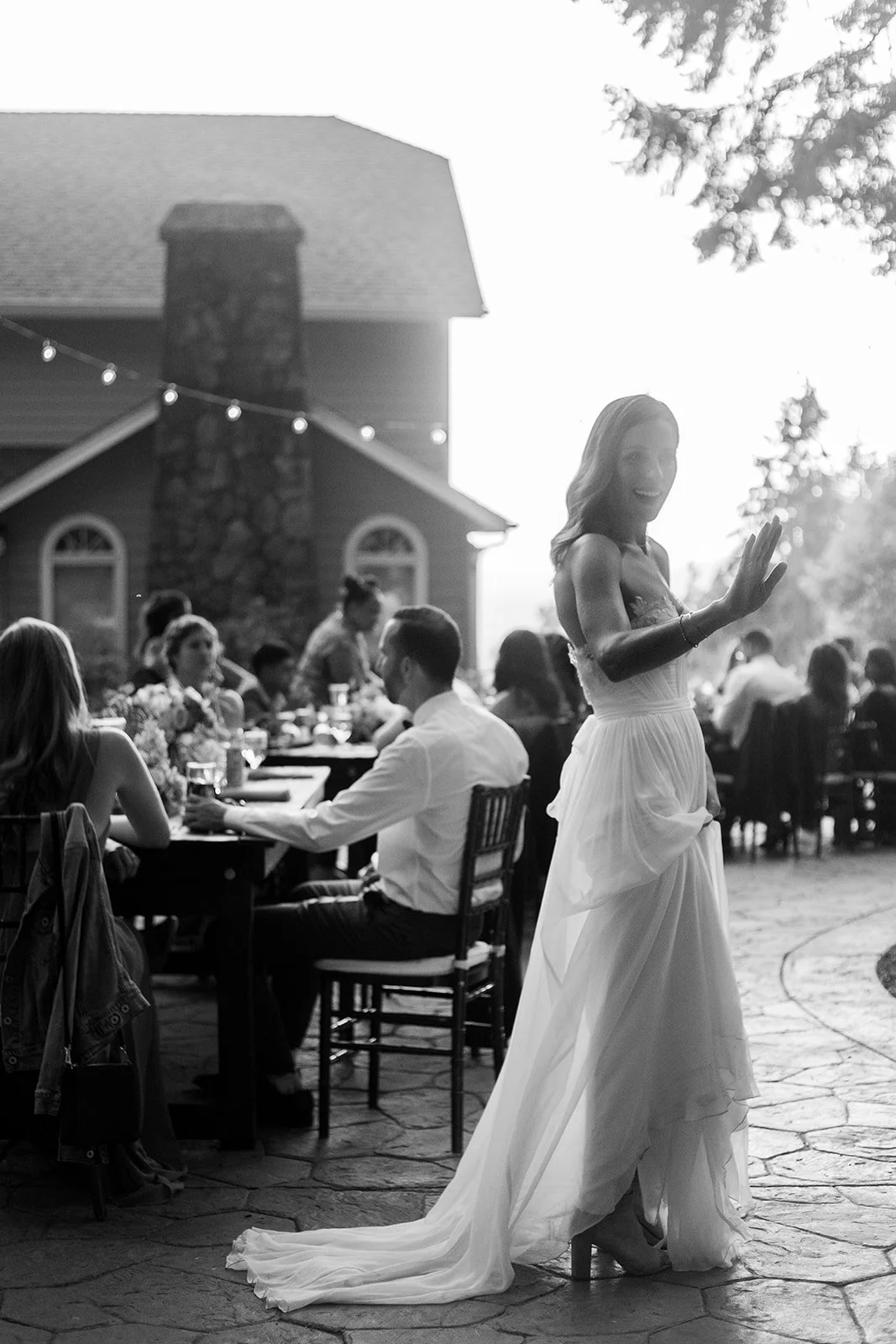 Woman in a white dress waving during an outdoor celebration beneath string lights.