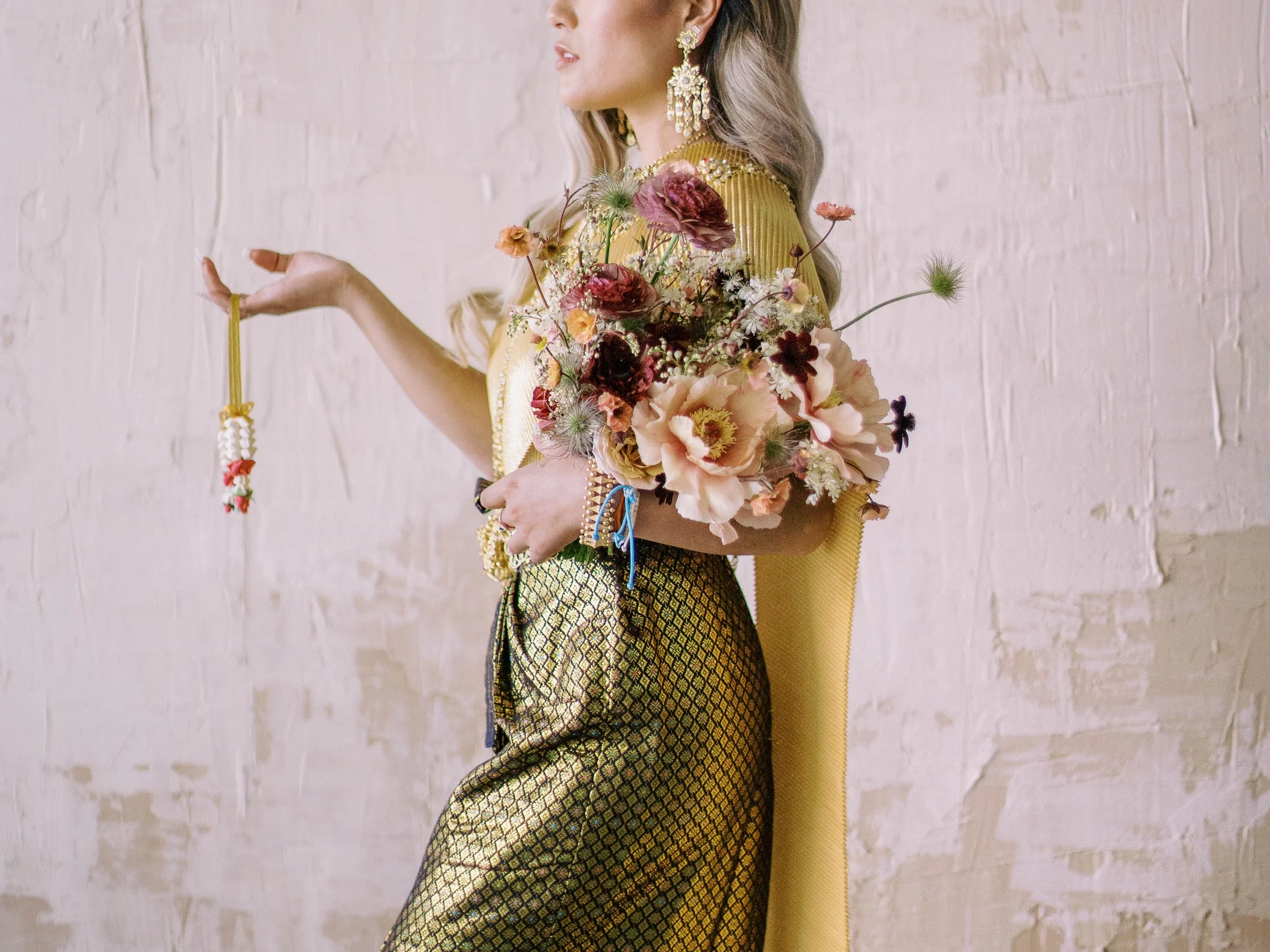 Woman in ornate gold and green traditional attire holding a colorful floral bouquet and floral garland.