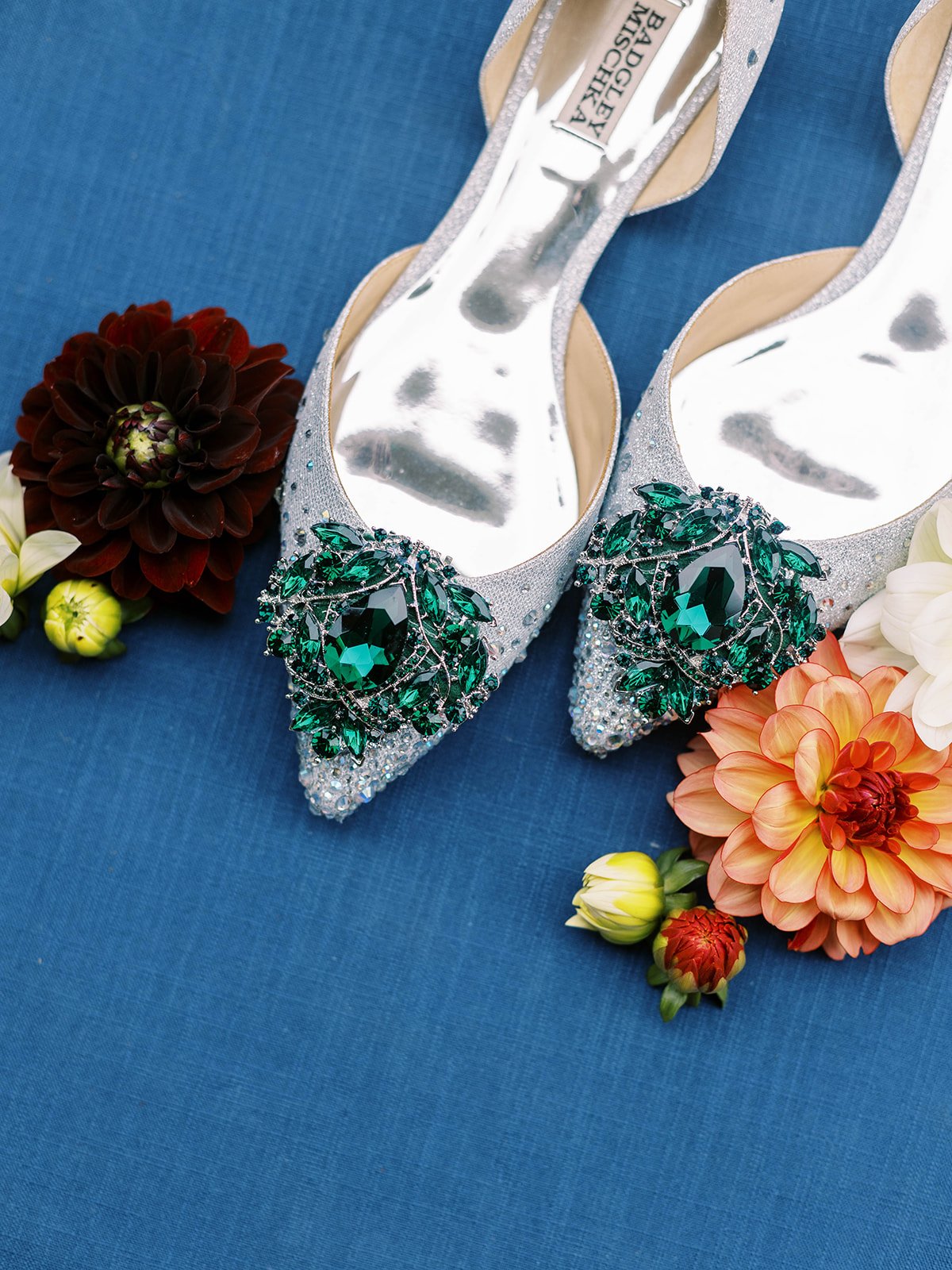 Emerald jeweled bridal shoes surrounded by orange and burgundy dahlias on a deep blue background.
