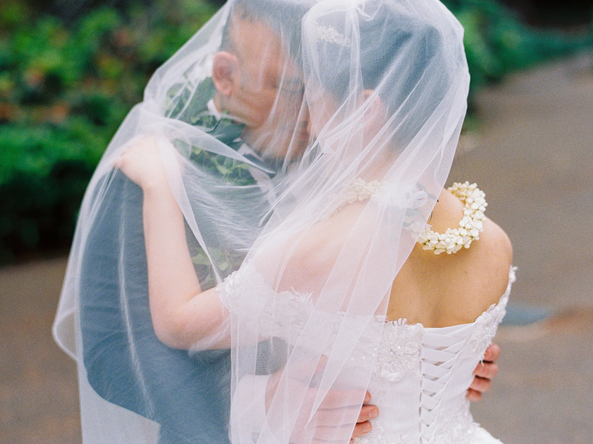 Bride and groom embrace beneath her veil, with the pua kalaunu lei visible across her shoulders