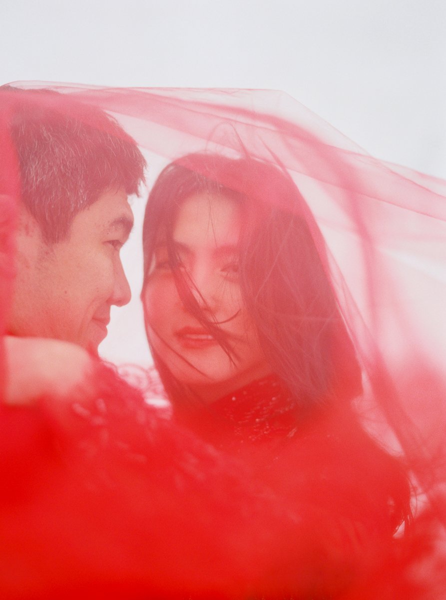 Artistic portrait of a couple wrapped in a flowing red veil, with the woman’s face softly visible through the fabric.