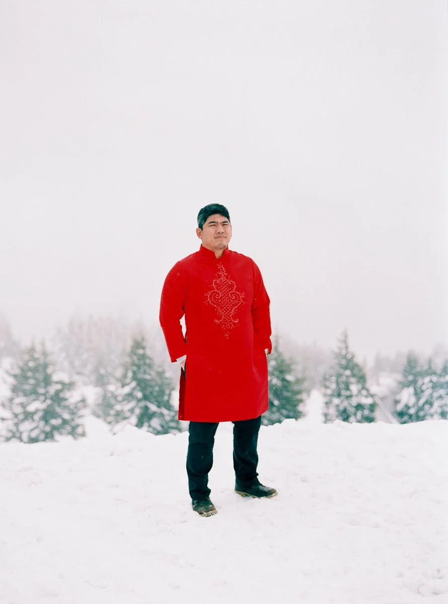 Man in a red embroidered áo dài standing alone in a snowy landscape with trees and fog in the background.