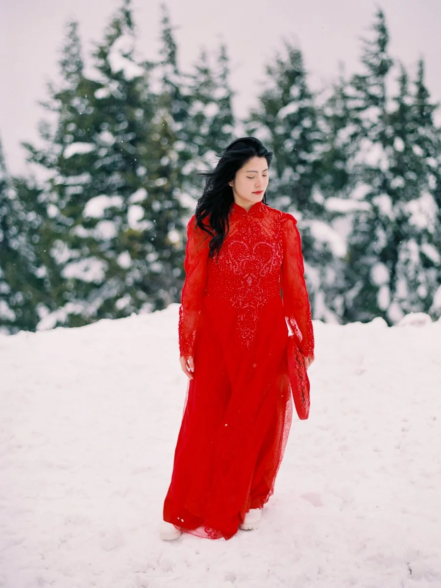 Woman in a vibrant red áo dài walking alone through a snowy landscape with evergreen trees in the background.