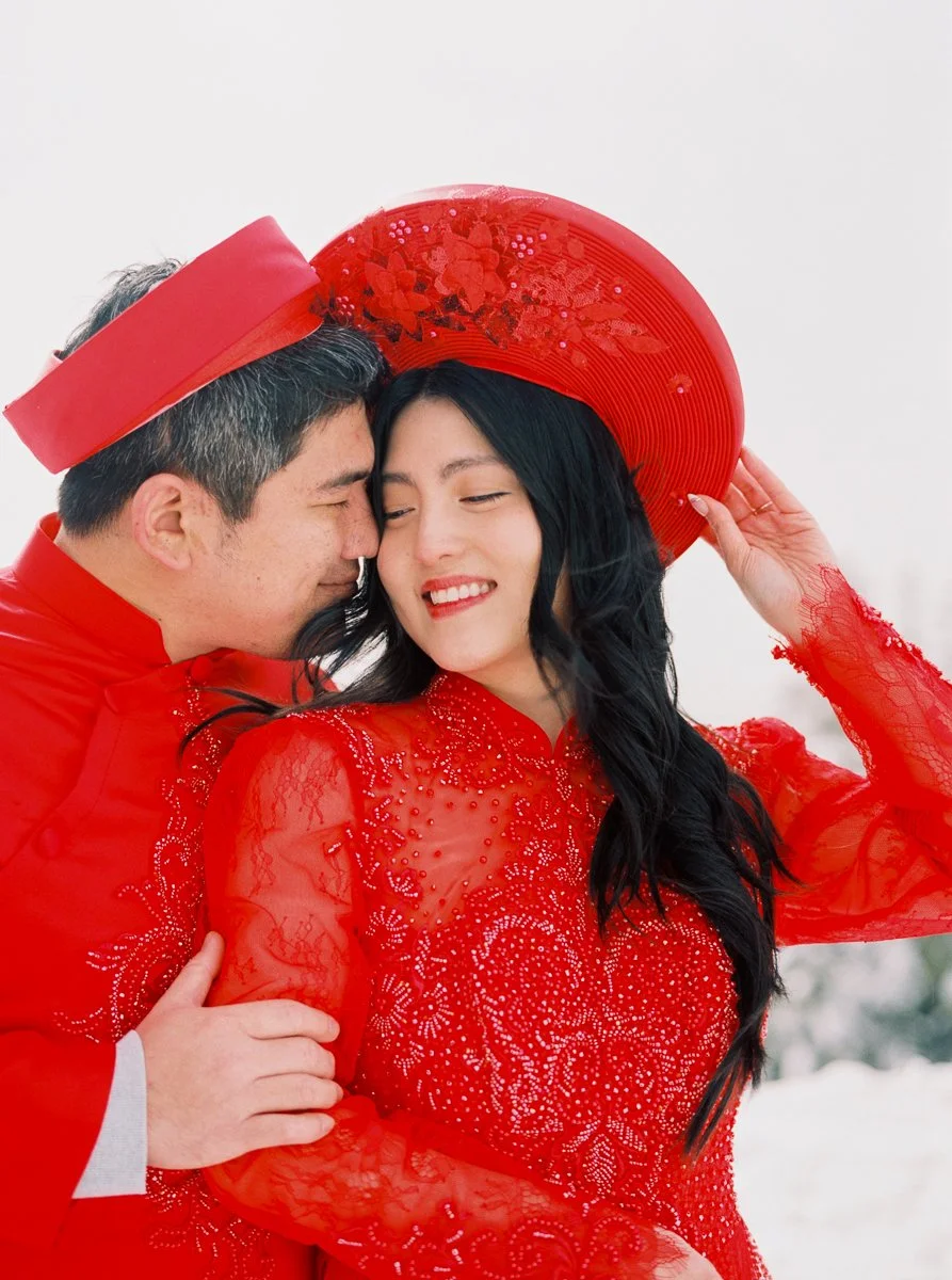 Man leans in toward a smiling woman as she adjusts her red khăn đóng; both dressed in matching traditional Vietnamese outfits.