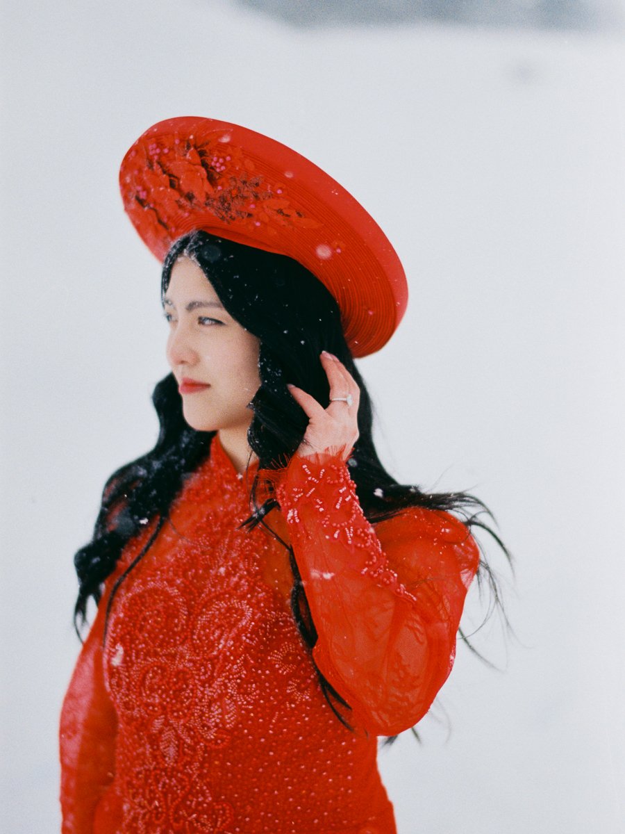 Woman wearing a red áo dài and matching khăn đóng looks off to the side, snowflakes landing in her long dark hair.