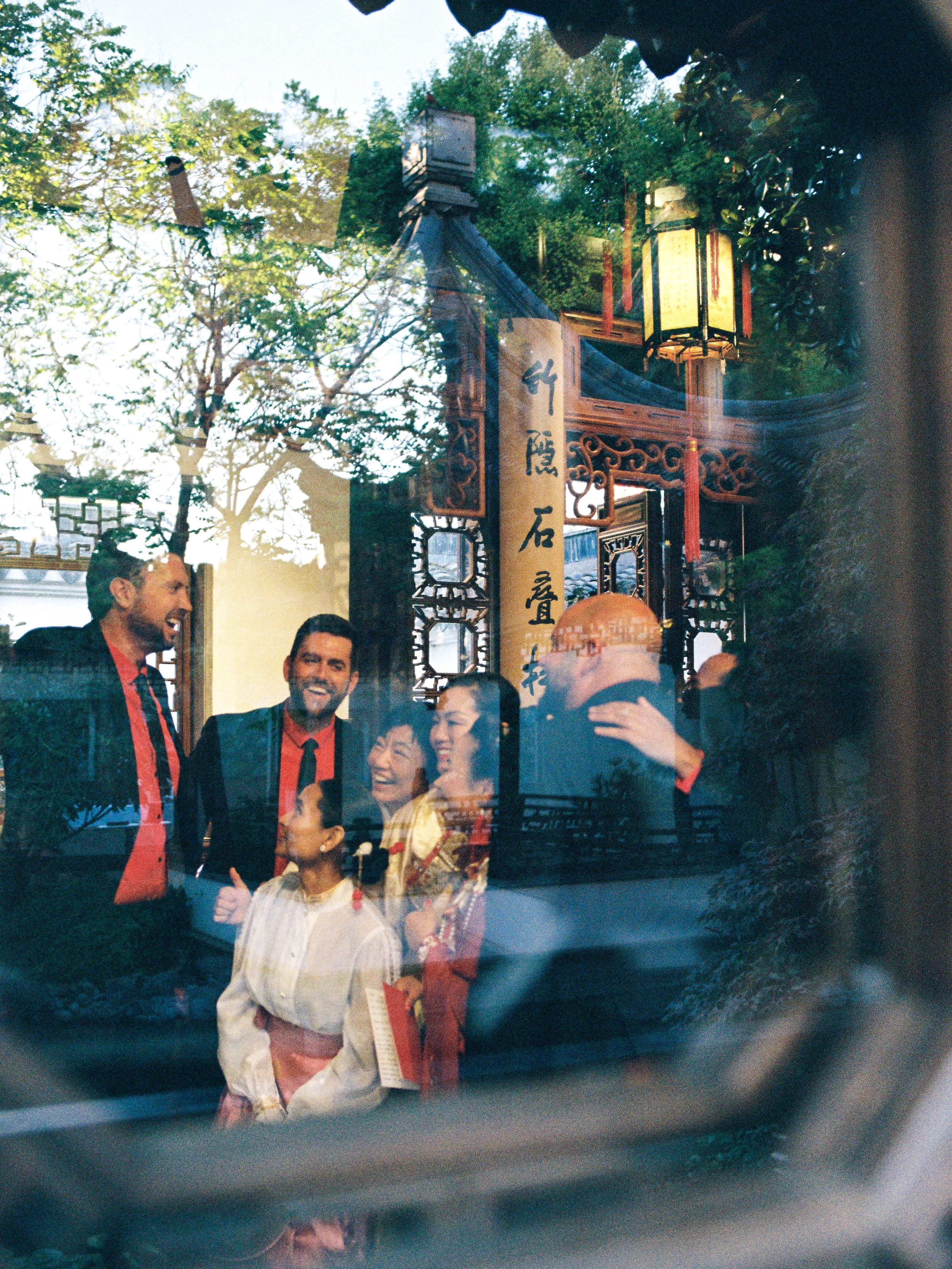 Wedding guests laughing together inside Lan Su Chinese Garden in Portland, seen through reflective glass and garden architecture during an intimate celebration.