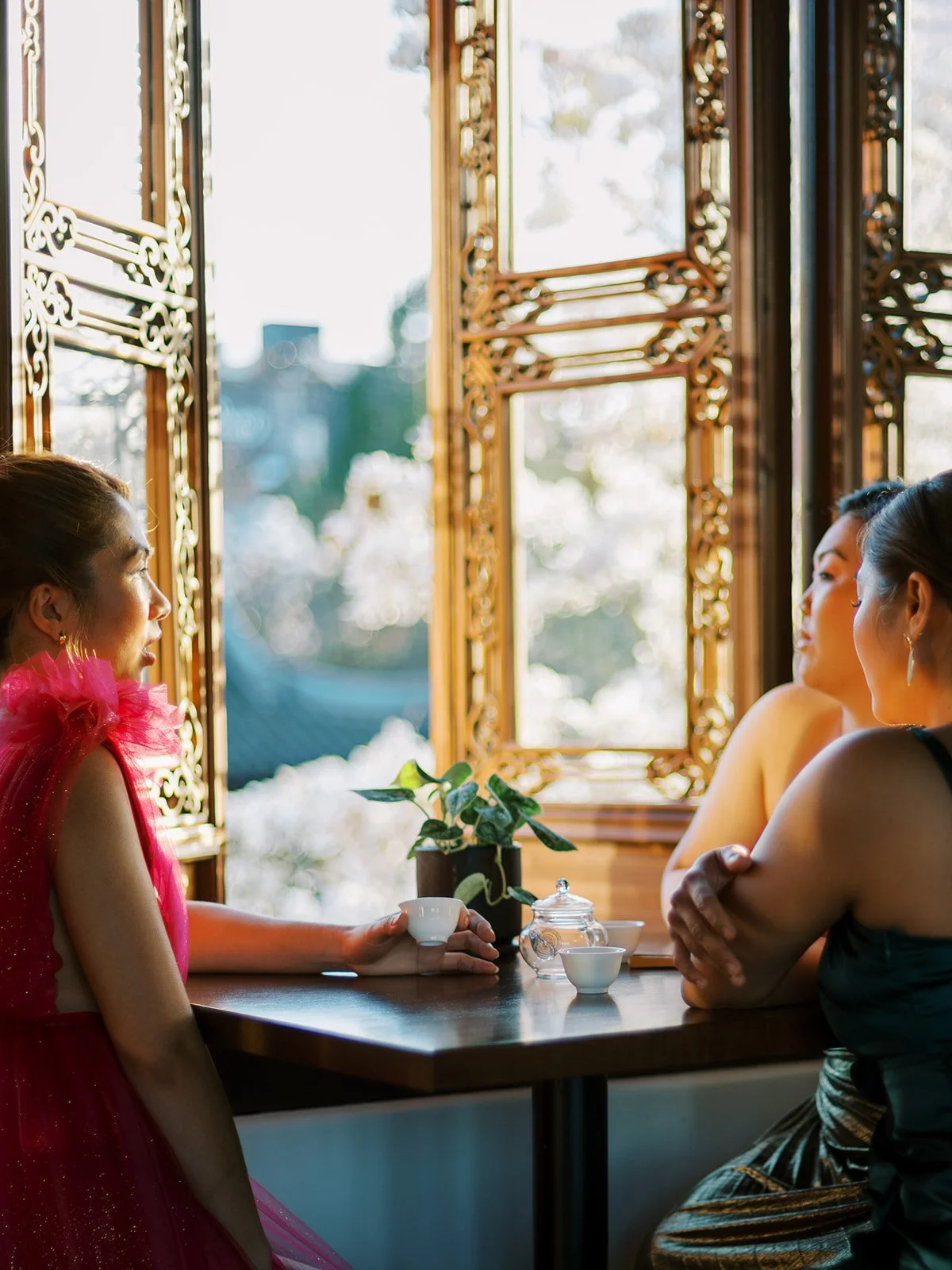 Women in couture gowns sharing tea by an ornate window during a Lan Su Chinese Garden bachelorette celebration in Portland.