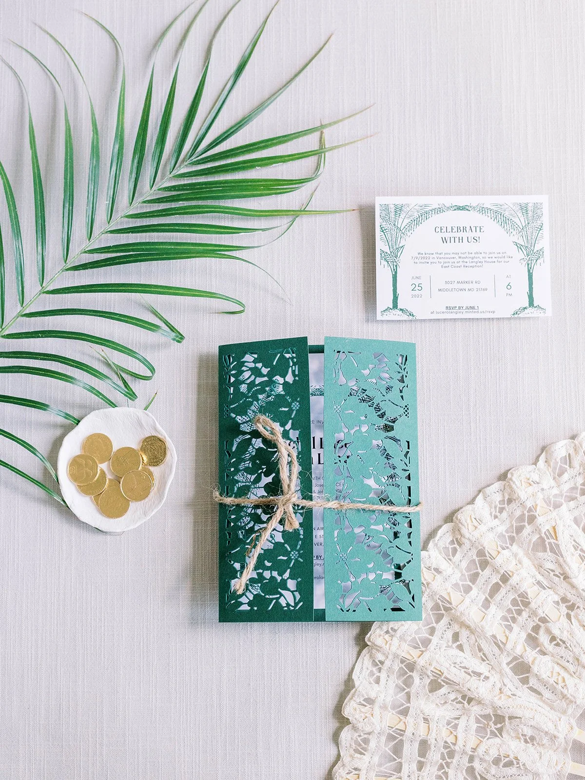 Flat lay of green laser-cut wedding invitations with palm leaf motifs, tied with twine and styled with lace, palm fronds, and gold coins.