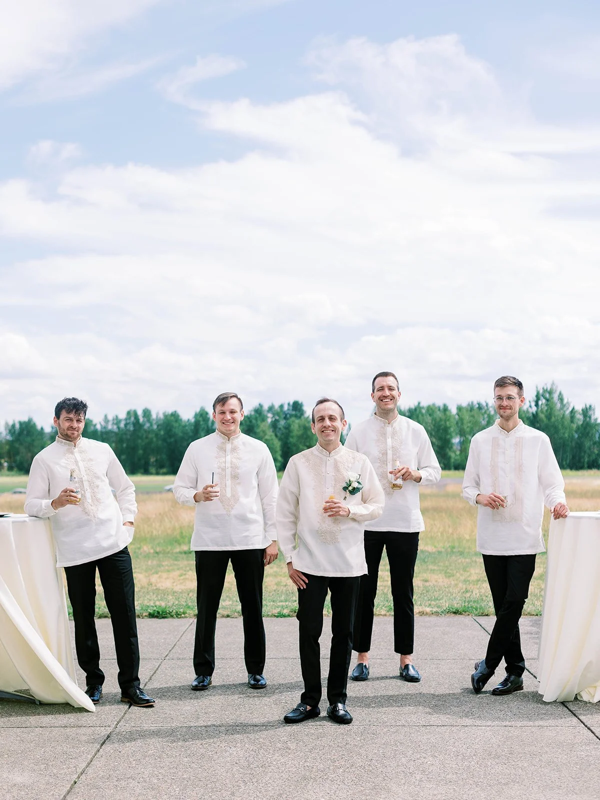 Wedding party wearing traditional Barong Tagalog shirts gathered outdoors at Pearson Air Museum, holding drinks and smiling together.