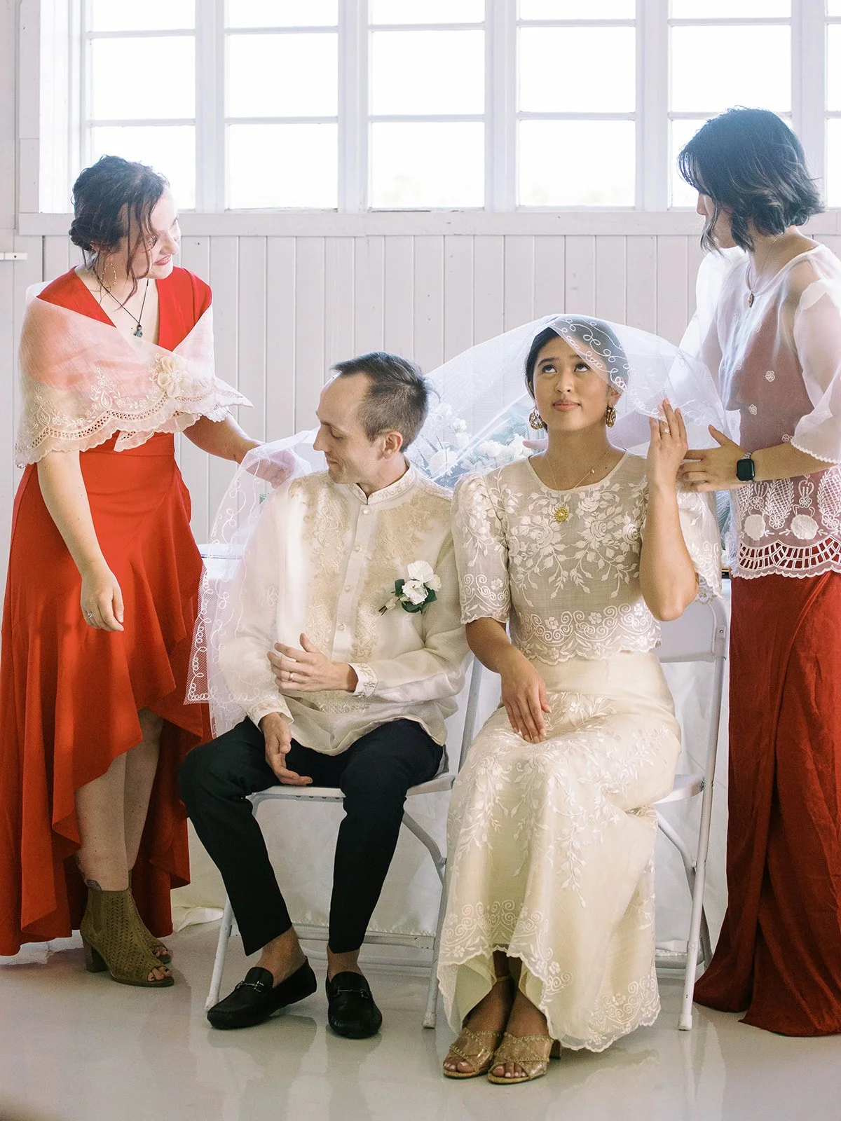 Bride and groom seated together as friends adjust the veil during a wedding ceremony inside the Pearson Air Museum hangar.