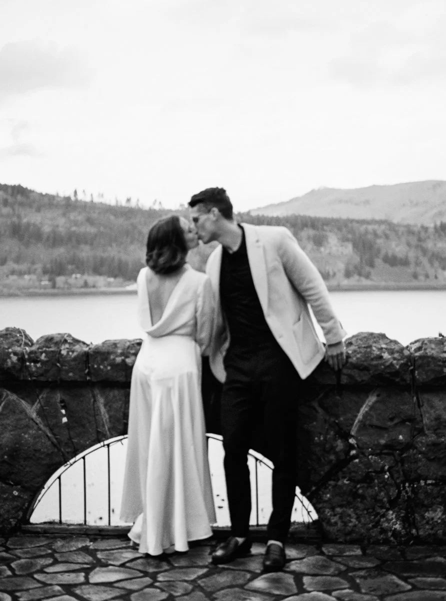 Black and white film image of the couple sharing a kiss at a stone overlook above the Columbia River Gorge.