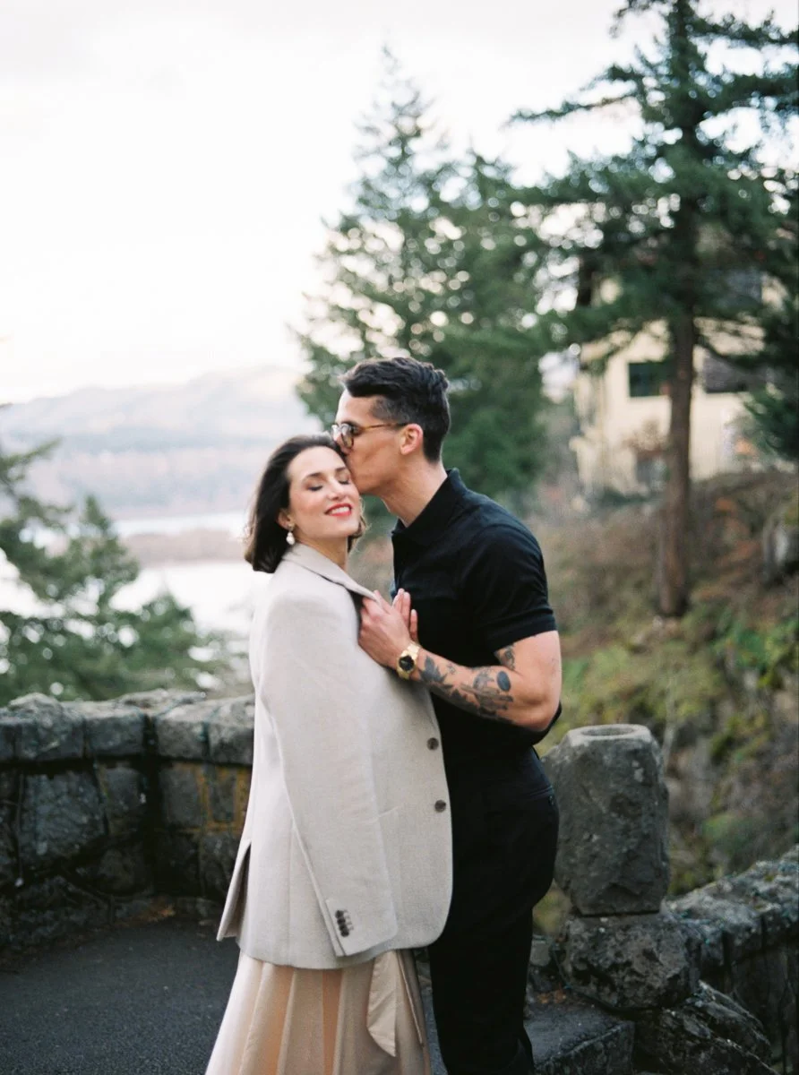 Groom kissing the bride’s temple at a stone overlook above the Columbia River Gorge, evergreen trees behind them, photographed on film.