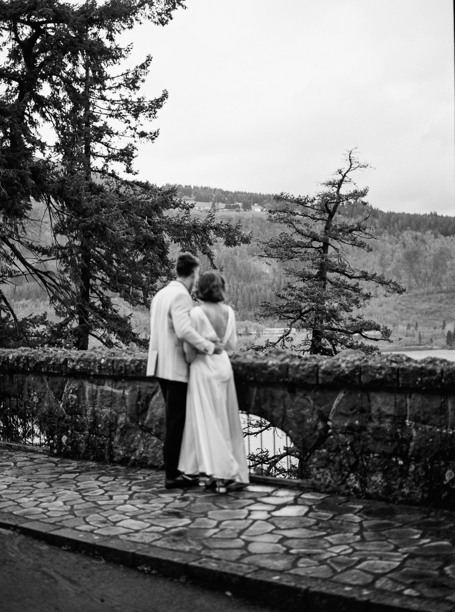 Black and white film photograph of the couple standing together at a stone overlook, looking out across the Columbia River Gorge.