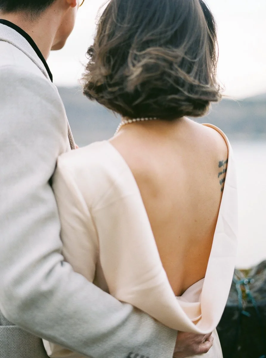 Close view of the bride’s open-back dress and pearl necklace as the couple stands together at a stone wall, captured on film.