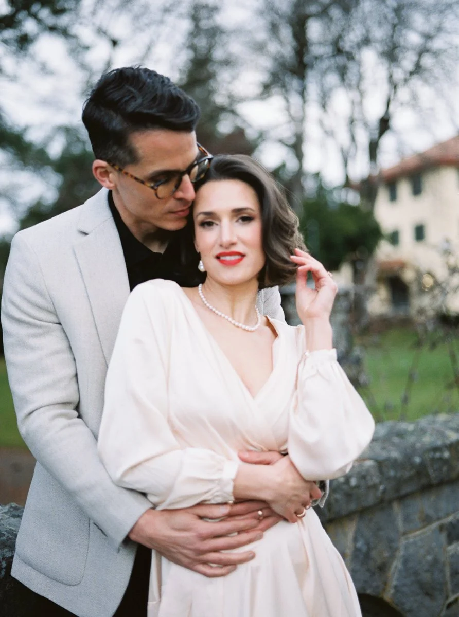 Color film portrait of the groom kissing the bride’s cheek as she looks toward the camera, framed by soft greenery.