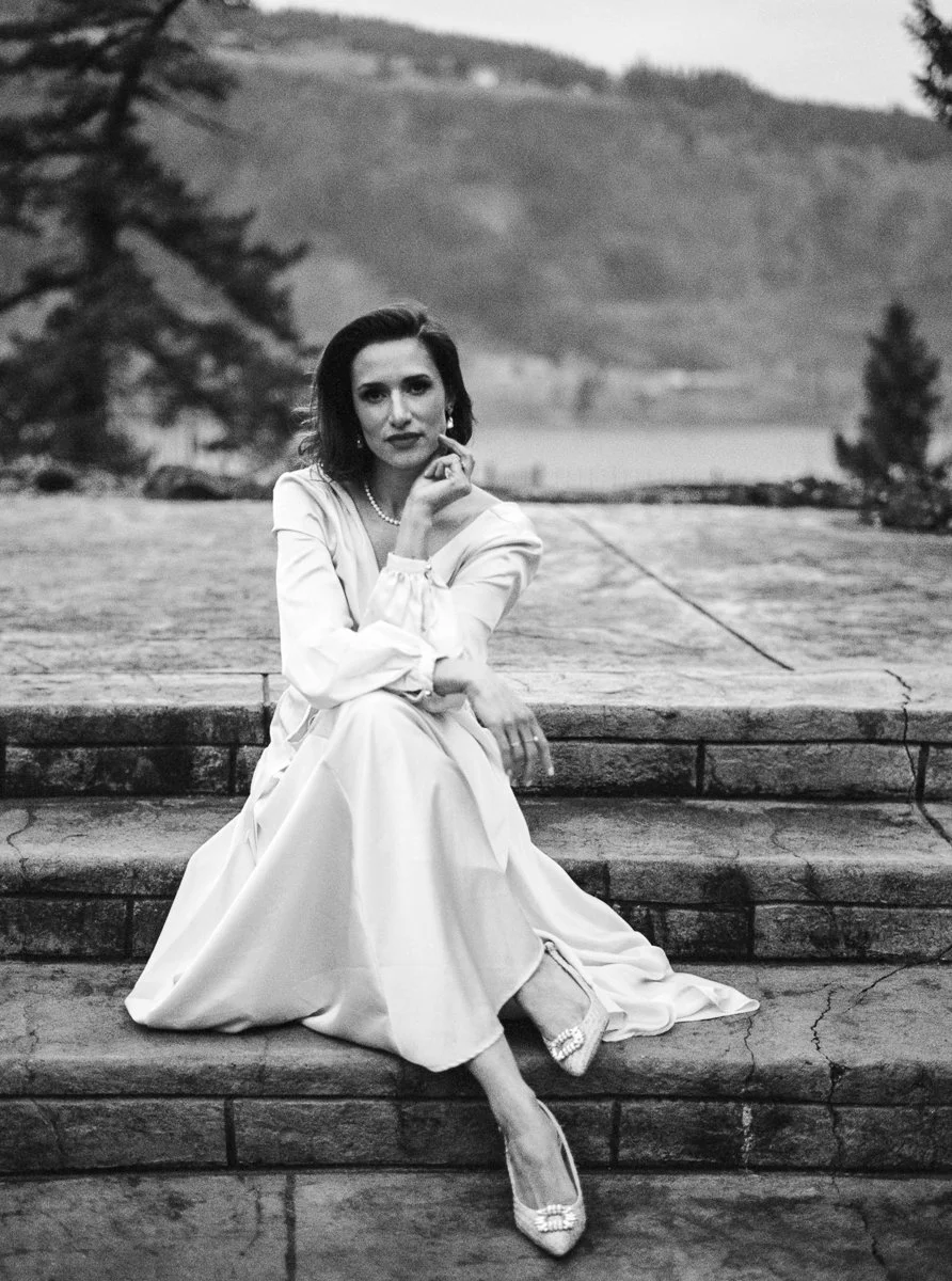 Black and white film portrait of the bride seated on stone steps, silk dress draped around her, framed by the Columbia River Gorge.