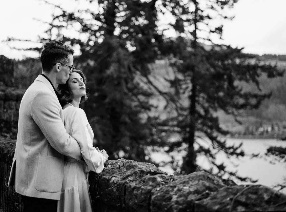 Couple embracing at a stone overlook above the Columbia River Gorge, evergreen trees in the background, photographed on black and white film.