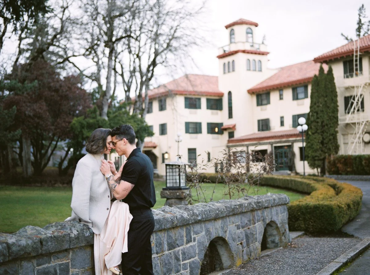 Couple standing close on a stone bridge at the Columbia Gorge Hotel, framed by historic architecture and winter trees, photographed on film.