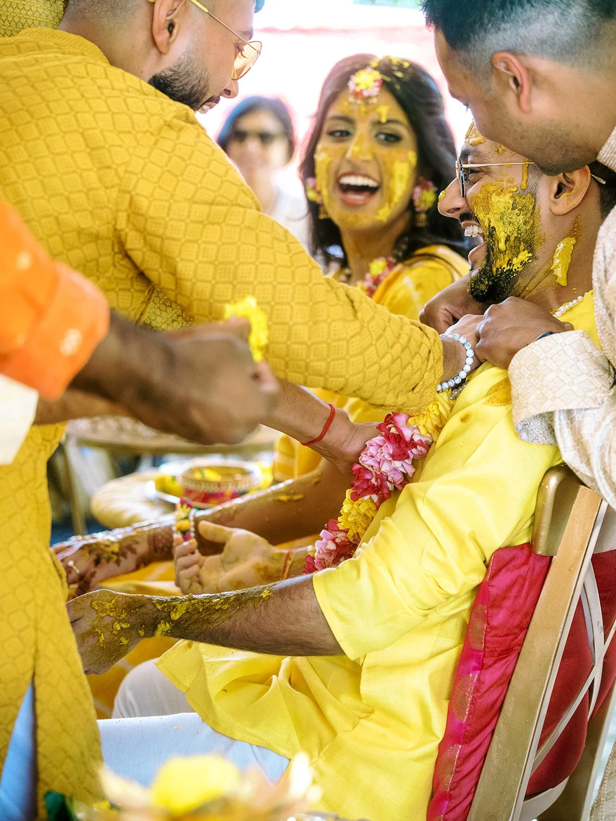 Family members applying turmeric paste to groom in yellow kurta during lively Haldi celebration.