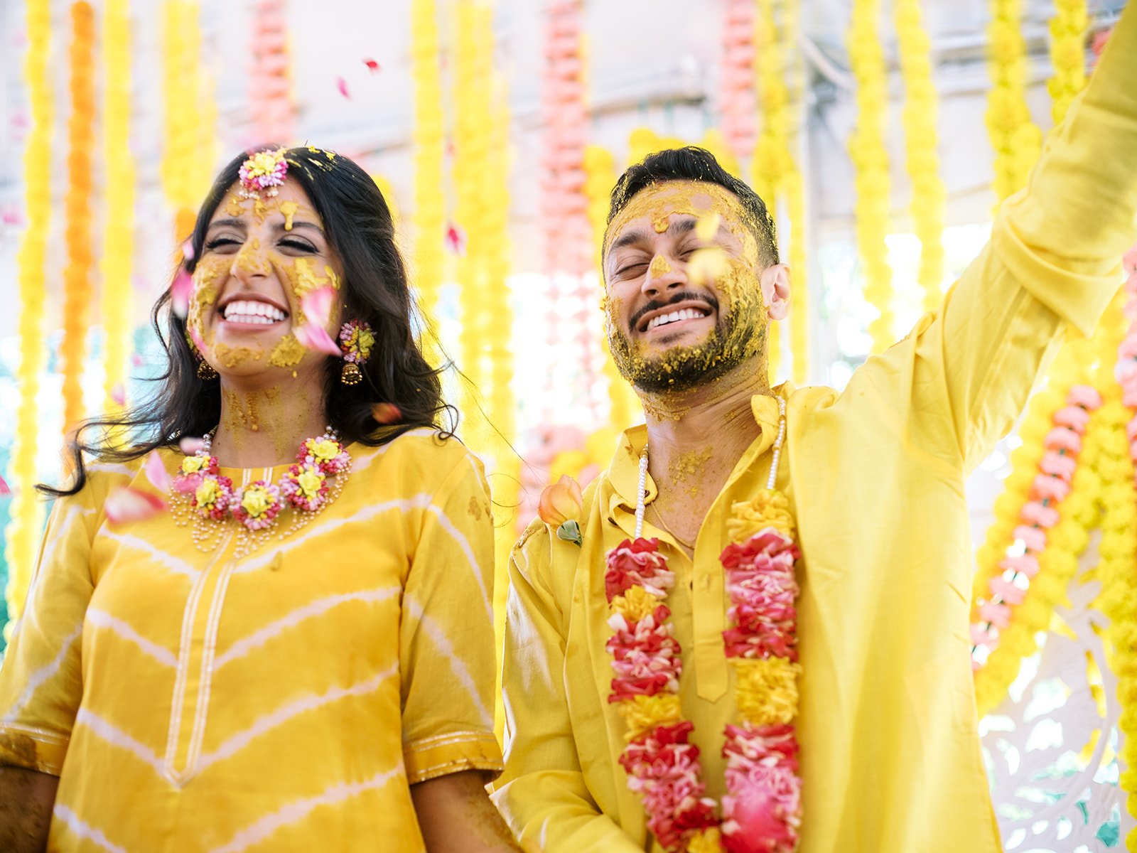 Bride and groom in yellow outfits covered in turmeric, smiling as flower petals fall around them.
