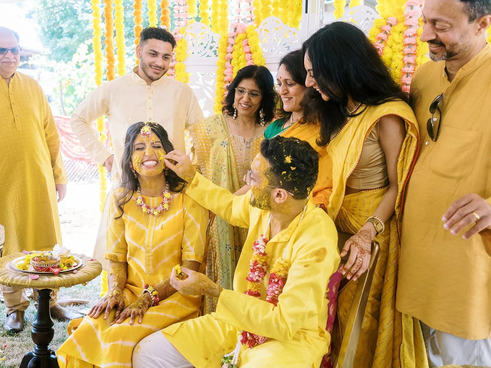 Bride seated in yellow dress as groom applies turmeric paste, surrounded by family in festive attire.