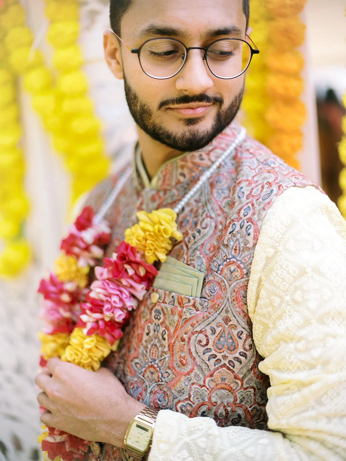 Groom in embroidered vest and cream kurta holding pink and yellow flower garland at Haldi ceremony.